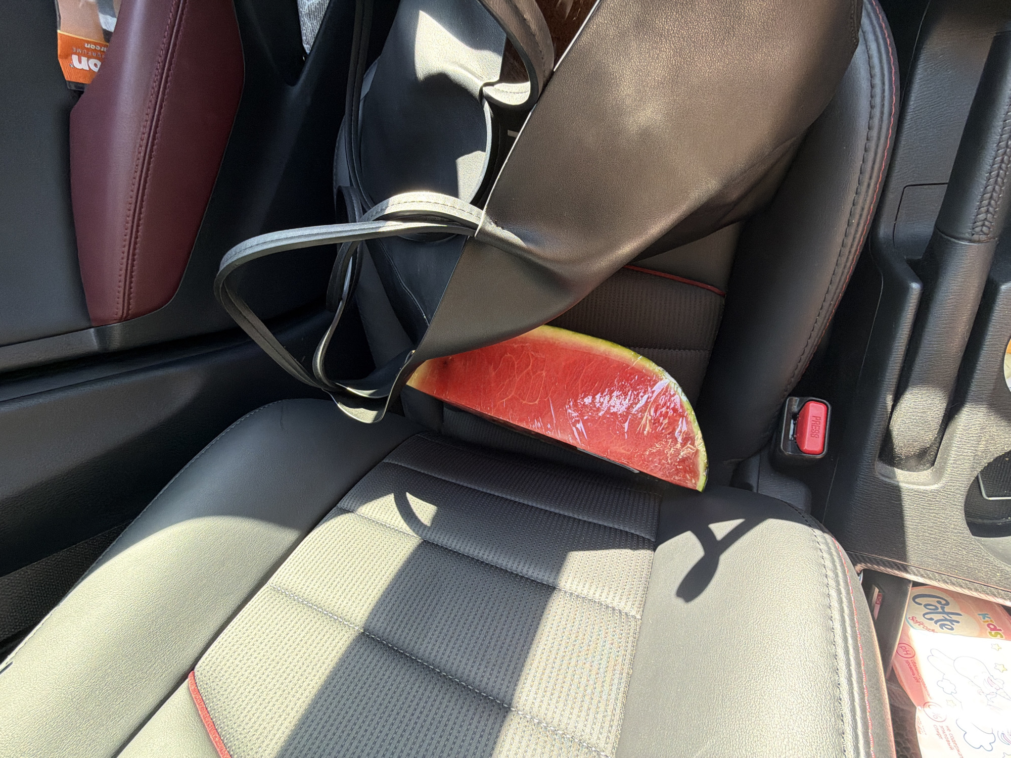 A slice of watermelon wrapped in plastic sits in the passenger seat of a car. A black bag is draped over the seat, partially covering the watermelon. The car has black leather seats with red stitching. A box of "Cotte Kids" soft care tissues is visible in the lower right corner of the image. The image was taken inside a car, presumably in the city of Chaikovsky, Russia.