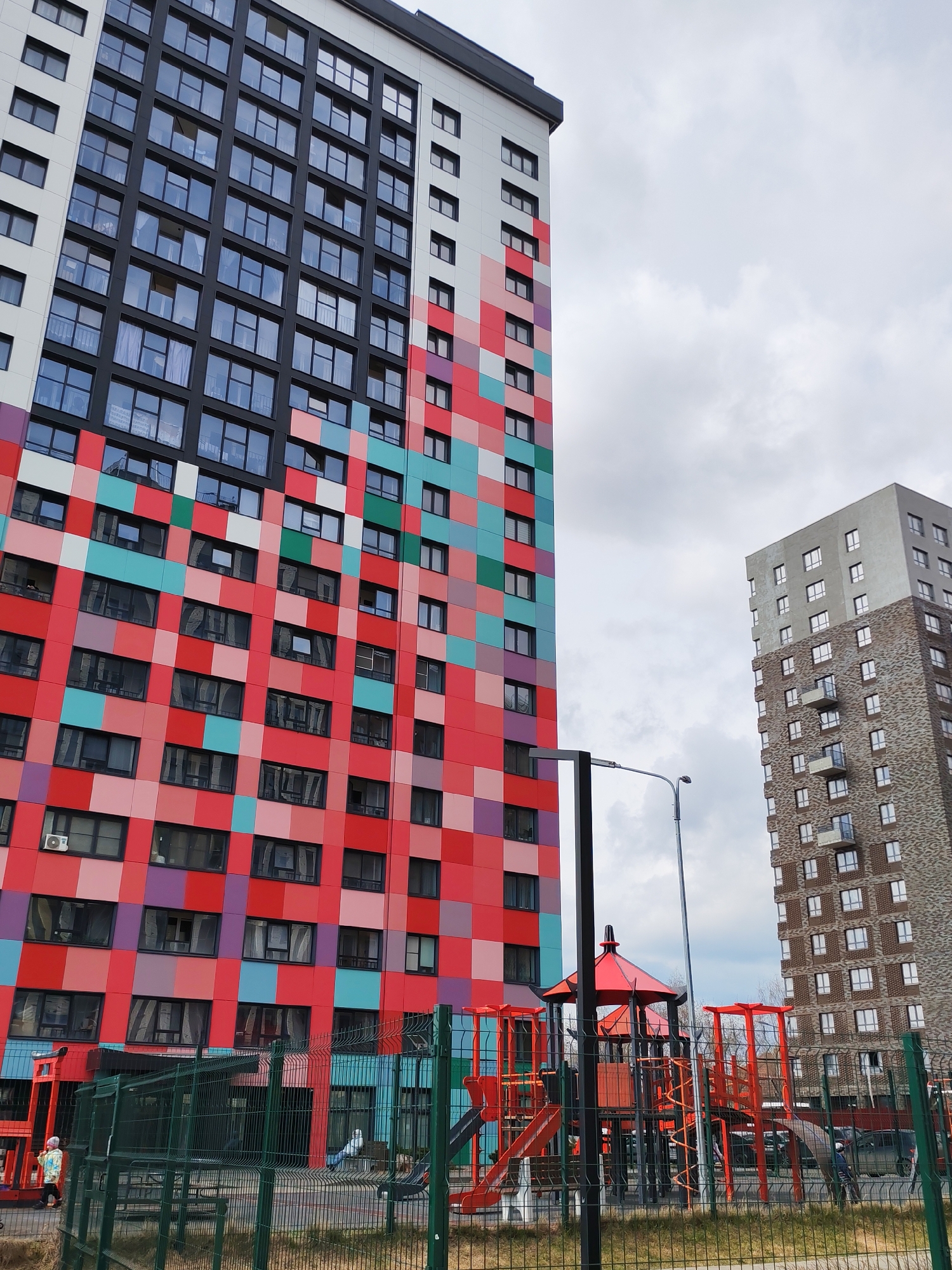 The photograph captures a scene outside a tall apartment building in Izhevsk, Russia. The building's facade is characterized by a vibrant, colorful pattern of red, pink, turquoise, and purple squares. The upper section of the building is black, white, and grey. Adjacent to the building is a playground, partially obscured by a green metal fence. The playground is painted in shades of red and orange. In the background stands another building, constructed with brown and grey bricks, and less vibrantly colored. The sky is overcast with a grey cloud cover. A street light stands prominently in the foreground. A person is partially visible near the fence.