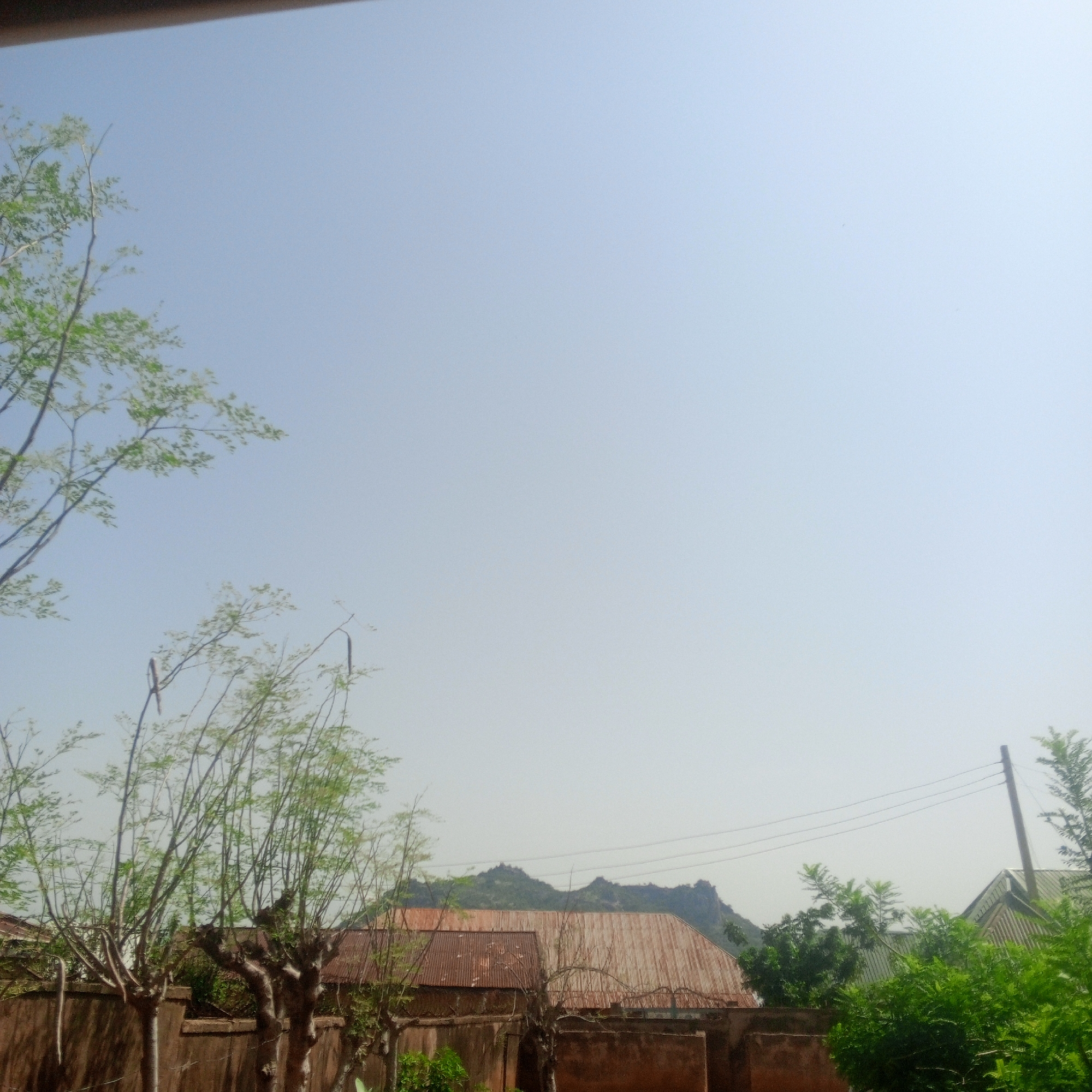 The image shows a residential area in Zinna, Nigeria, under a clear, pale blue sky. In the foreground, several trees with sparse green leaves and thin branches are visible, some appearing to be Moringa trees. A brown, textured wall runs horizontally across the lower portion of the frame, with some vegetation growing around it. Behind the wall, several buildings with corrugated metal roofs are present. One building has a rusted, reddish-brown roof, while another has a lighter colored corrugated roof. In the background, a rocky, green mountain range is visible. A utility pole with wires stretches across the upper right portion of the image, near some trees with dense green foliage. The overall impression is of a sunny day in a rural or semi-rural setting within Zinna, Nigeria.