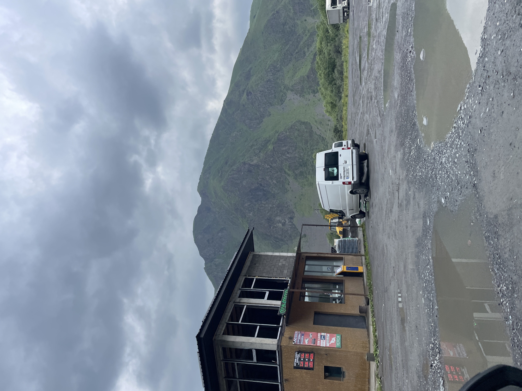 The image shows a building with a sign that says "SIM CARD" and "XEROX" in Stepantsminda, Georgia. A white van is parked in front of the building. In the background, there is a large green mountain and a cloudy sky. The ground in front of the building is wet and has puddles. There is also a yellow excavator parked behind the van. The scene appears to be a rest stop or a service area along a road.