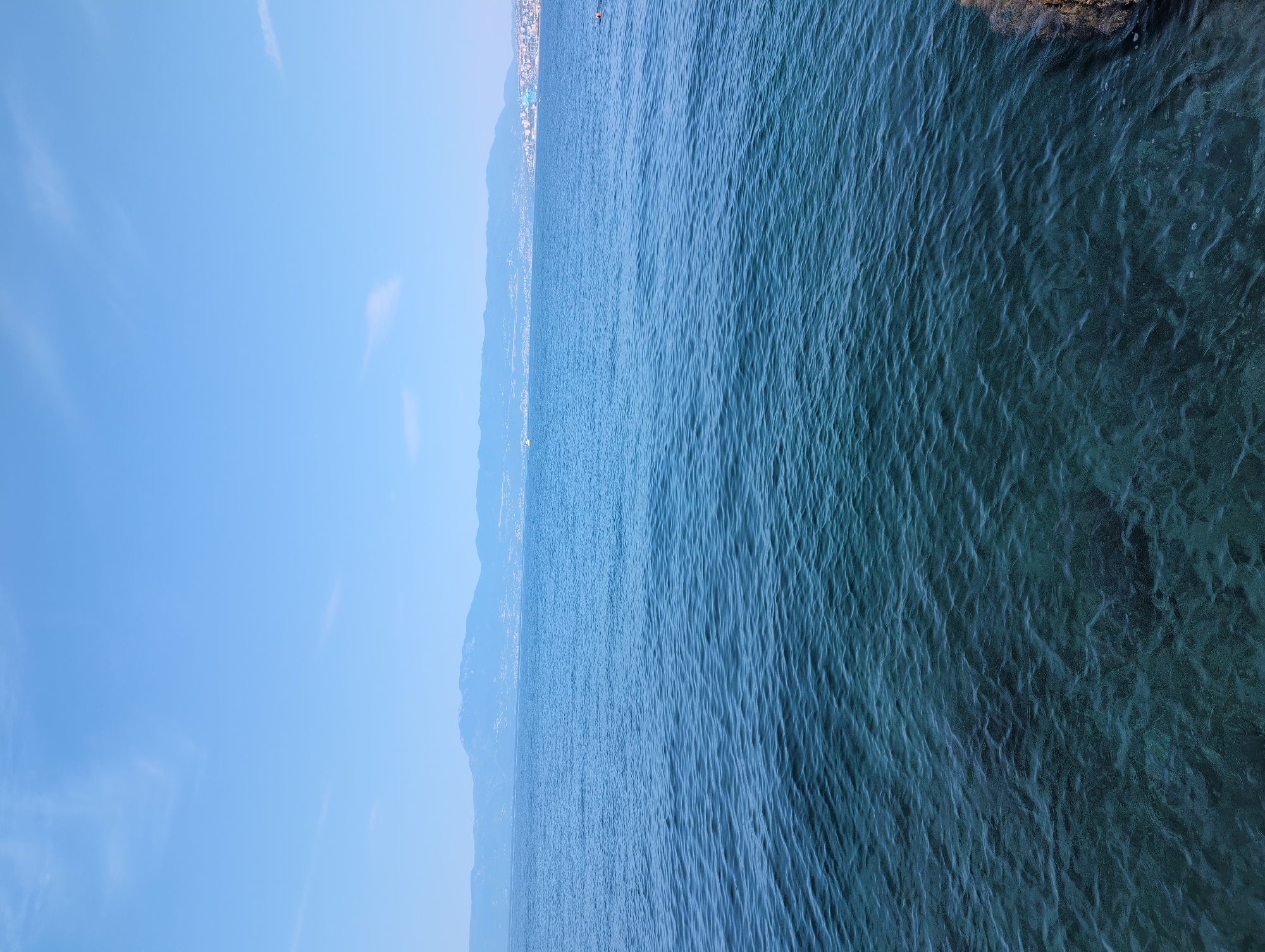 The image depicts a serene seascape looking out towards a distant, hazy coastline under a clear blue sky. The foreground is dominated by the deep blue, slightly rippled surface of the sea, with the seabed visible in the shallower, clearer water near the bottom right, revealing rocks and possibly submerged vegetation. A rocky outcrop is visible in the bottom right corner. In the mid-ground, the water stretches towards a sprawling coastline dotted with buildings, suggesting a city or town. The background features rolling hills and mountains that fade into the atmospheric haze. The sky is a bright, cloudless blue, with a few wispy clouds visible towards the top left. The overall impression is one of a calm, sunlit day at the coast.