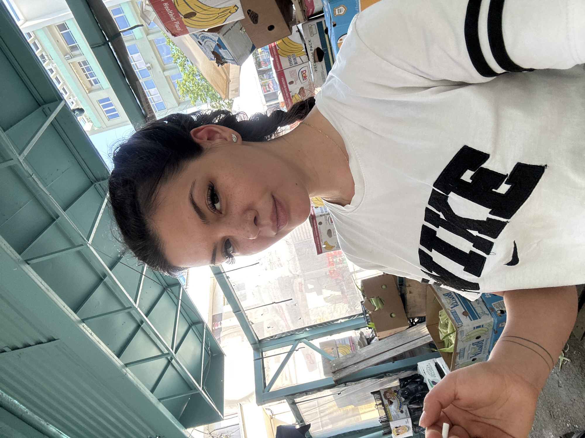 A young woman with dark hair braided into a single plait is taking a selfie. She is wearing a white Nike t-shirt with black stripes on the sleeves. She is holding a cigarette in her left hand and has a tattoo on her left forearm. She is outdoors, possibly at a market, with boxes of fruit visible in the background. Buildings are visible in the background, suggesting an urban environment. The image appears to have been taken in Sliven, Bulgaria, based on the context provided.