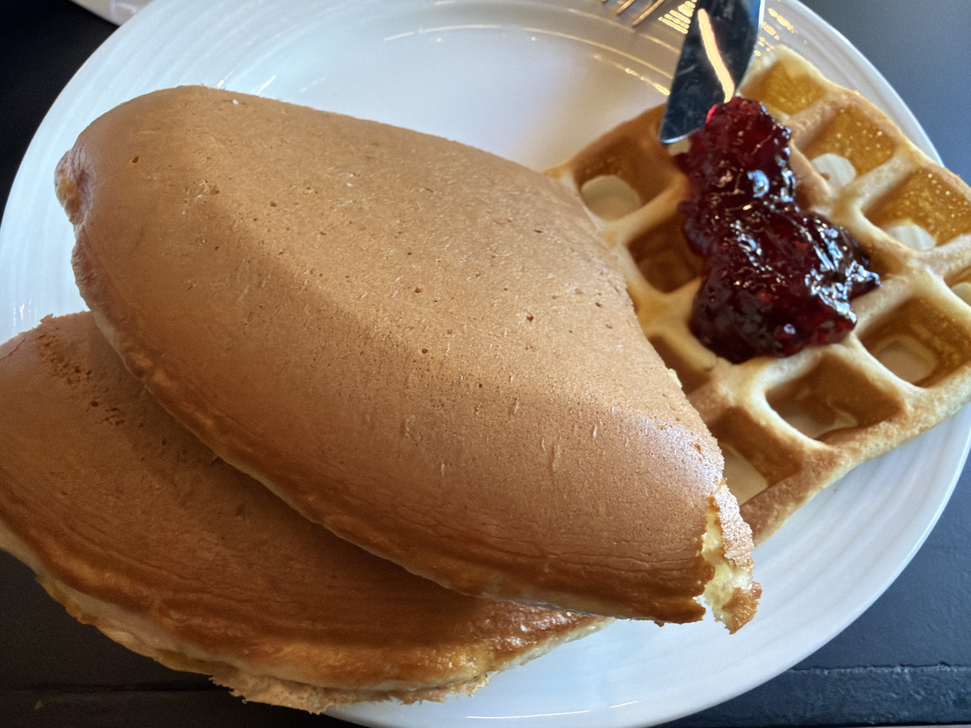 A close-up shot captures a breakfast plate featuring two thick, fluffy pancakes and a section of a golden-brown waffle. The pancakes, stacked one on top of the other, are light brown with a smooth, slightly porous texture. The top pancake shows a rounded, risen form, suggesting a light and airy consistency.
Adjacent to the pancakes, on the white, round plate, lies a piece of a waffle with a classic grid pattern. A generous dollop of dark red jam or fruit preserve, possibly raspberry or mixed berry, rests in the center of the waffle, its glossy surface reflecting the light. The jam appears chunky with visible fruit pieces. A silver utensil, likely a fork or knife, is partially visible at the top right, with its handle resting near the edge of the waffle and jam.
The plate itself is white and subtly textured, with a slight rim. The background is a dark, undefined surface, possibly a table, providing a contrast to the bright food and plate. The lighting is bright and even, indicating daytime, possibly morning given the nature of the meal. The overall scene suggests a prepared breakfast, perhaps in a restaurant or cafe in Tbilisi, Georgia. No people or visible text are present in the image.