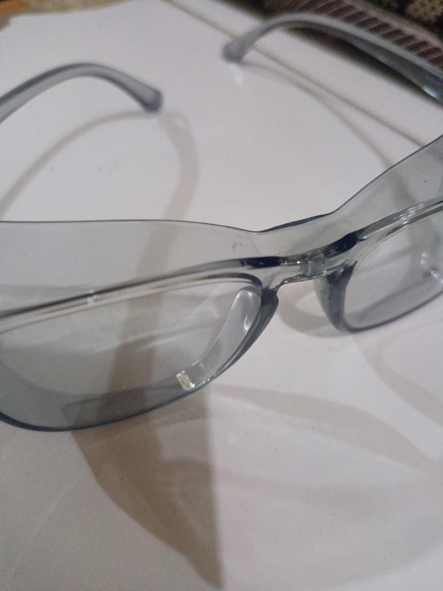 The image shows a close-up, slightly blurry shot of a pair of clear safety glasses, possibly tinted, resting on a light-colored, textured surface. The glasses are made of plastic and have a wraparound design. The left lens and part of the frame are in focus, while the right lens is out of focus. Shadows are cast on the surface by the glasses, indicating a light source from the top right. There are some minor smudges and imperfections visible on the lenses. The background shows a suggestion of a pattern, possibly fabric, at the top of the image. There are no people, actions, or specific environmental details that would indicate a location in the City of Medan, Indonesia.