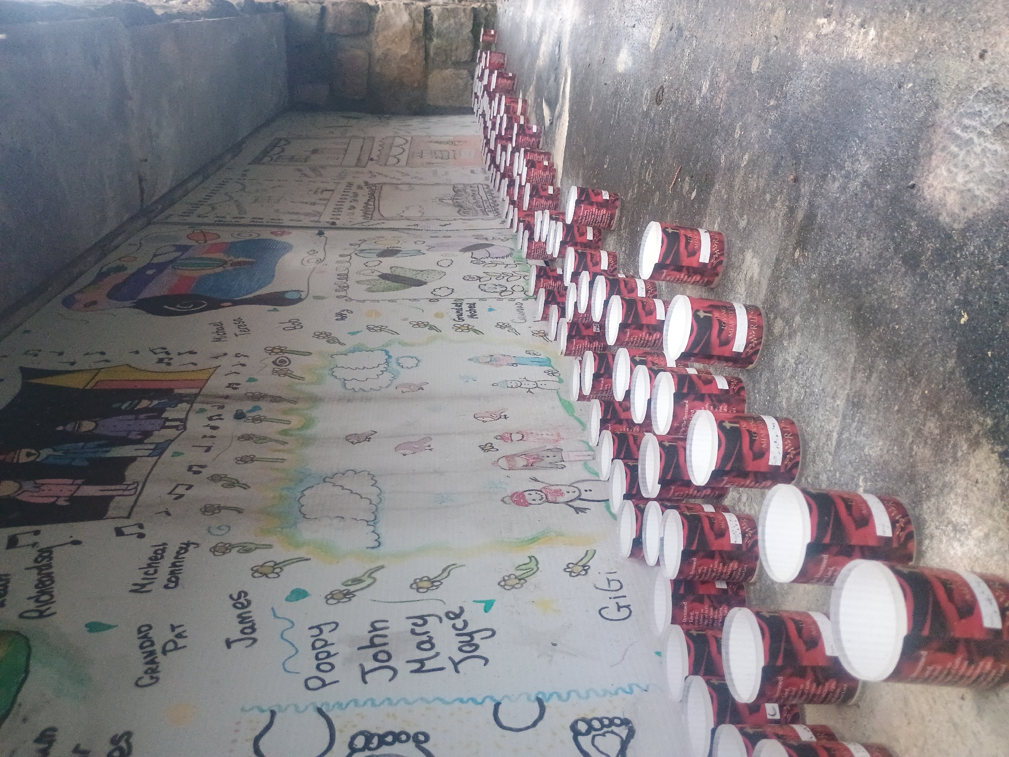 In an outdoor, possibly urban, environment, a stone wall with painted murals forms the backdrop for a scene featuring rows of white-topped cylindrical objects, likely candles or luminaries, arranged on a concrete surface. The wall displays a colorful mural with abstract shapes, figures, and text, including names like "Grandad Pat," "Michael Conroy," "James," "Poppy," "John," "Mary," and "Joyce." Further along the wall, smaller drawings of snowmen, birds, and flowers are visible. The cylindrical objects are predominantly red with black and white patterned labels, some of which appear to have religious text and imagery, including the words "Deceased Lord" and "In Loving Memory." The objects are organized in a dense, staggered arrangement, extending from the foreground into the middle ground of the image. The ground surface is textured concrete, with some debris and variations in color. Overhead, a concrete structure or overhang is visible, casting shadows on the scene. The lighting suggests it is daytime, with natural light illuminating the area.