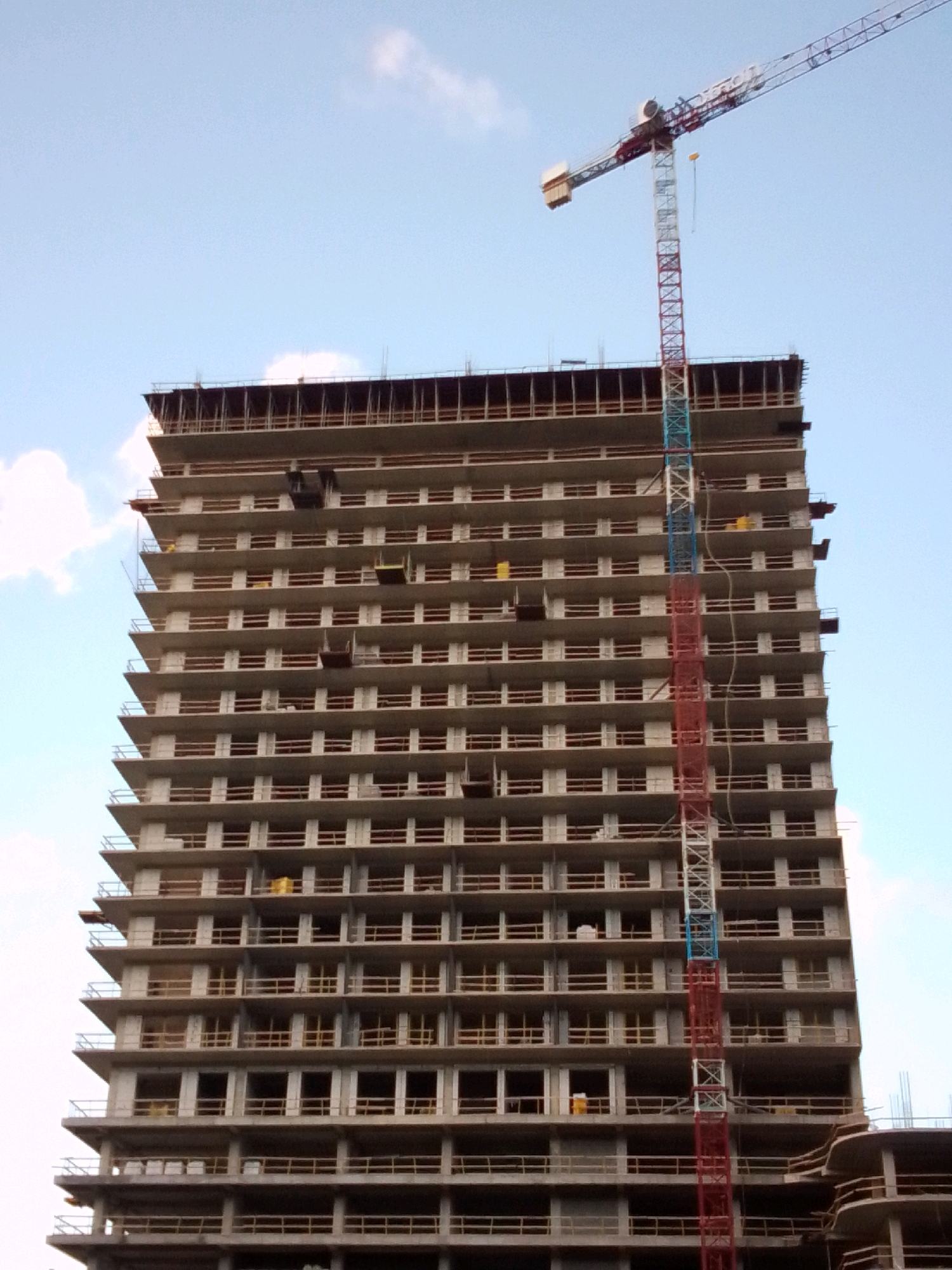 A construction crane with a red, white, and blue segmented tower stands beside a multi-story building under construction. The building, made of concrete, has numerous open floors with visible scaffolding and protective barriers. The crane's horizontal arm extends above the building, and its counterweight is visible on the left side. The sky above is a light blue with scattered white clouds. The scene is an outdoor construction site, likely in the city of Tbilisi, Georgia, given the building style and typical construction cranes seen in developing urban areas. No people are visible in the image.
