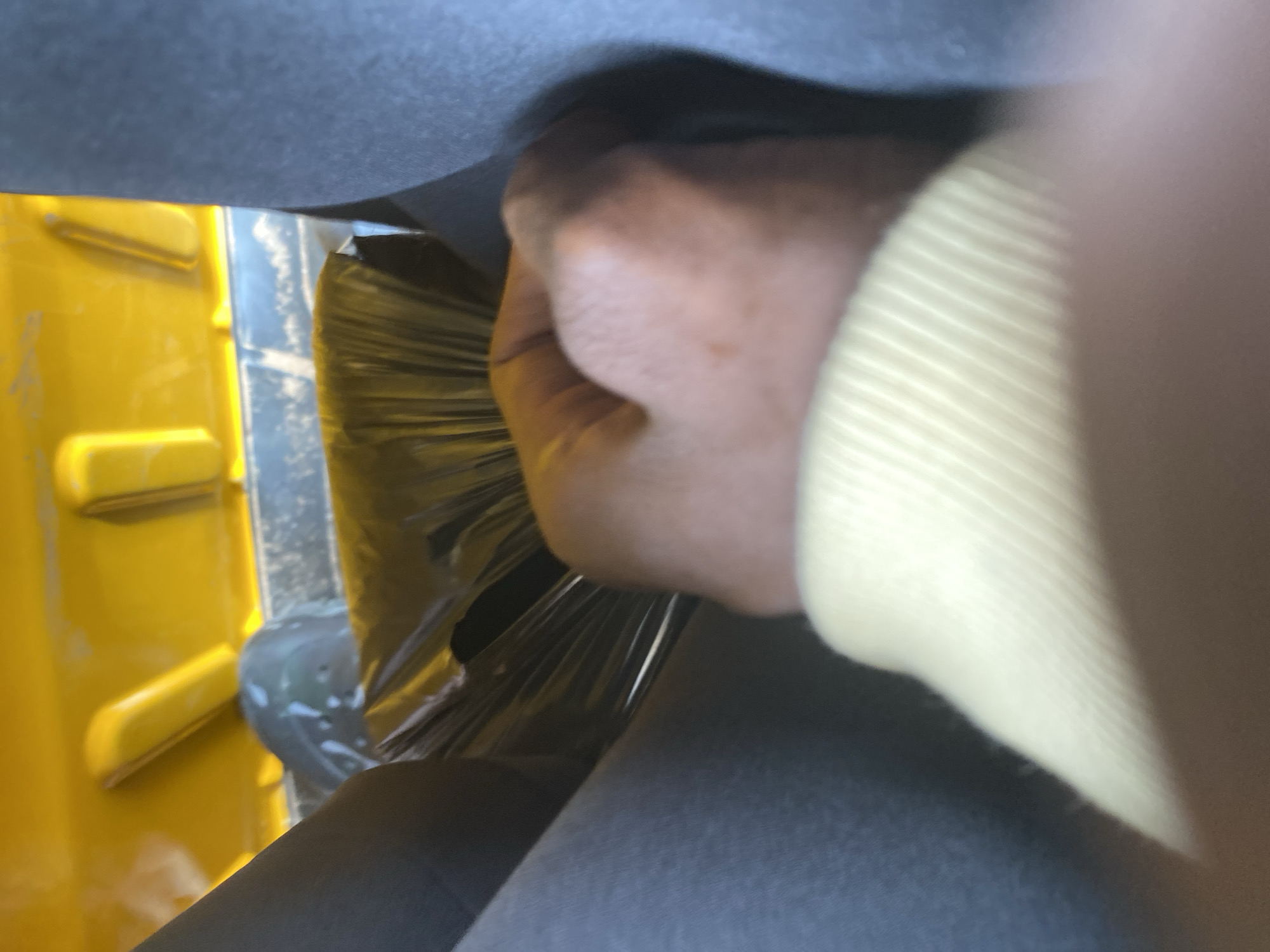 A hand is seen holding a brush made of fine, transparent plastic bristles, which are fanned out. The brush appears to be cleaning a part of a yellow, rectangular object that has raised horizontal lines. The hand is holding the brush with a closed fist, and the palm is facing upwards. A portion of a light yellow garment is visible on the right side of the image, possibly worn by the person whose hand is shown. The background consists of dark gray material, suggesting an indoor setting or the interior of a vehicle. The image is taken at a close-up angle, focusing on the interaction between the hand, brush, and the yellow object. There are no visible text elements in the image.