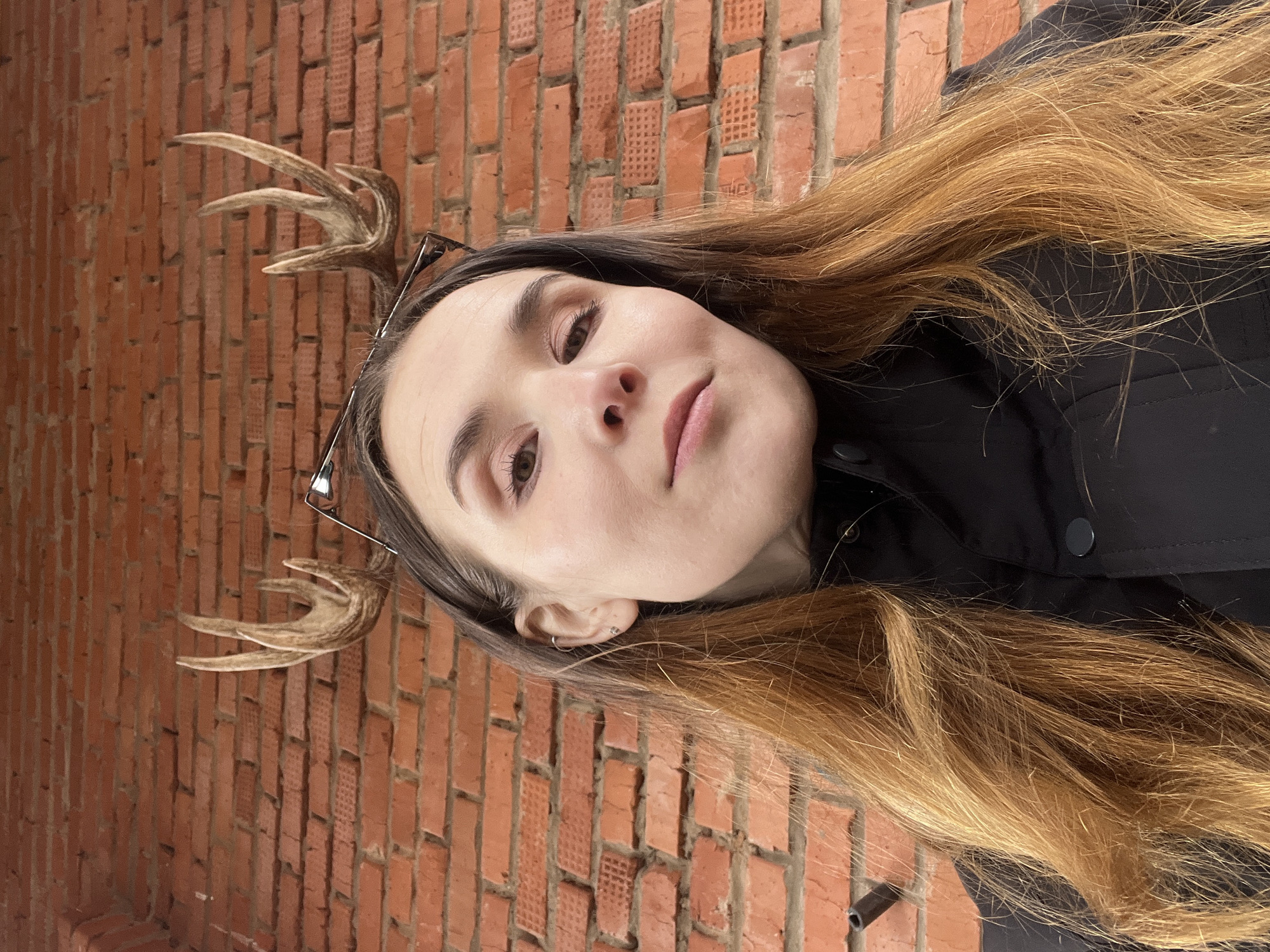 A young woman with brown and blonde hair stands in front of a brick wall. She is wearing a black jacket and glasses, which have small deer antlers attached. The location is unknown and cannot be determined to be Votkinsk, Russia.
