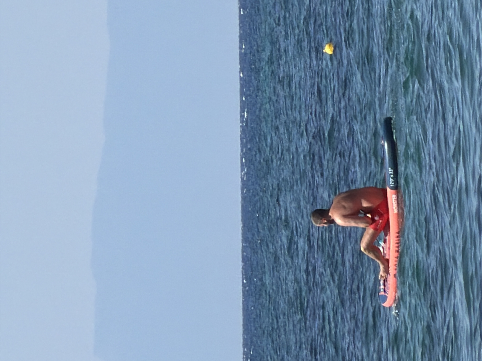 A person is sitting on an orange paddleboard in the sea off the coast of Nea Iraklia, Greece. The person is facing to the left, with their legs bent and their arms resting on the board. They are wearing red shorts. The paddleboard has text printed on it, including "AQUA MARINA" and "MONSTER". The sea is choppy and a yellow buoy is visible in the distance. The sky is clear and blue, with a faint outline of hills or mountains on the horizon.