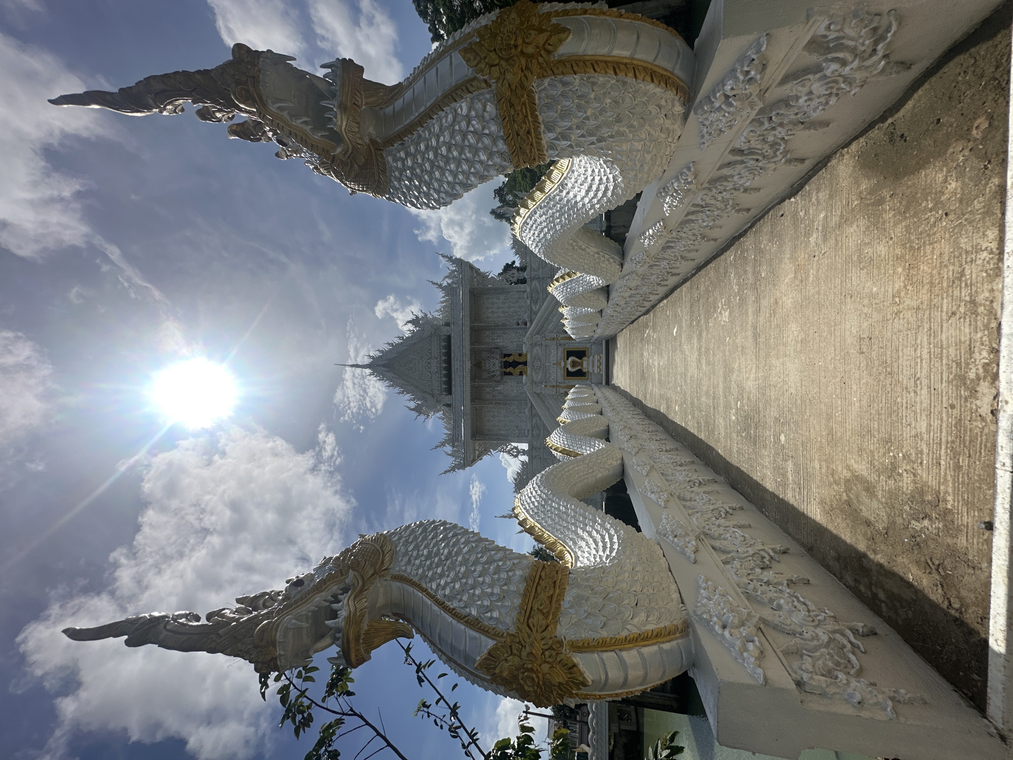 A wide-angle, low-angle shot captures a pathway leading towards a white temple with ornate architecture under a bright, sunny sky. Flanking the pathway are two large, white, serpent-like statues with gold accents, their heads rearing upwards. The pathway itself is made of concrete and is lined with decorative carvings on the sides of the serpent statues' bodies. The sun is a prominent feature, creating a lens flare in the upper center of the image, with scattered clouds in the blue sky. Green foliage is visible to the left of the frame. The overall scene suggests a spiritual or religious site in a tropical or subtropical environment. This location is Wat Phra Nang Sang in Thep Krasatti, Thailand.
