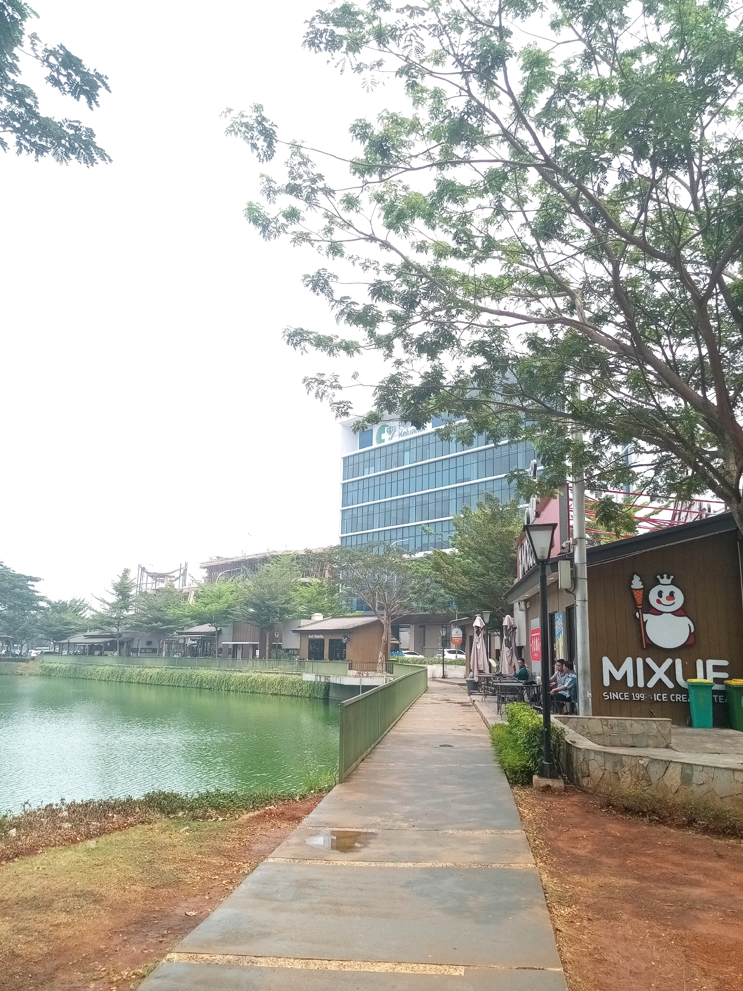 This is an eye-level, medium shot of a walkway beside a green body of water, with a row of shops and a tall building in the background, likely taken in Bekasi, Indonesia. The walkway is made of concrete with some puddles, and a low green fence runs along the water's edge. The water is a bright, vibrant green with a slight ripple. In the background, there are several low-rise buildings, appearing to be shops or restaurants, with tables and umbrellas set up outside. Behind these, a modern, multi-story glass building dominates the skyline. The sky is overcast, appearing bright white. Trees with green foliage are visible throughout the scene, with a large tree in the foreground on the right casting shadows. A sign with a cartoon snowman and the text "MIXUE" and "SINCE 1997 ICE CREAM & TEA" is prominently displayed on one of the shops. Two individuals are seated at a table outside one of the shops.