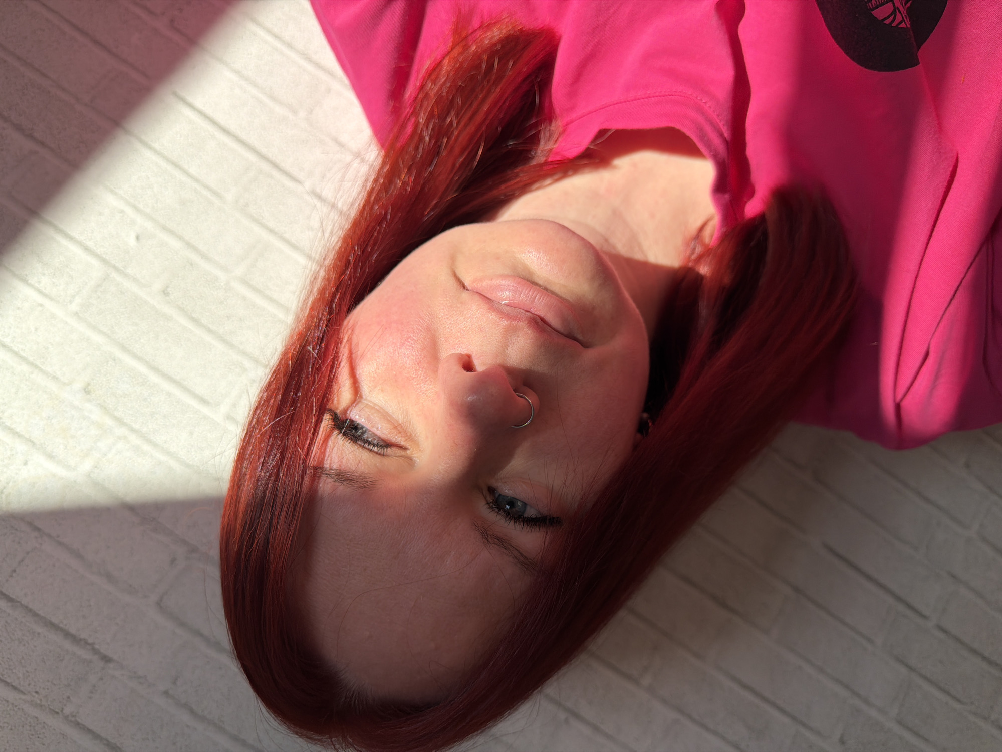 A Caucasian woman with shoulder-length red hair is lying on what appears to be a white brick or tiled surface, possibly a wall or floor. She is wearing a bright pink long-sleeved shirt or sweatshirt. She has a nose ring in her left nostril and appears to be looking slightly down and to the left. The light source is from the left, casting a shadow across half of her face. The image is rotated counter-clockwise, so the woman appears to be lying on her side. There is no indication of location.