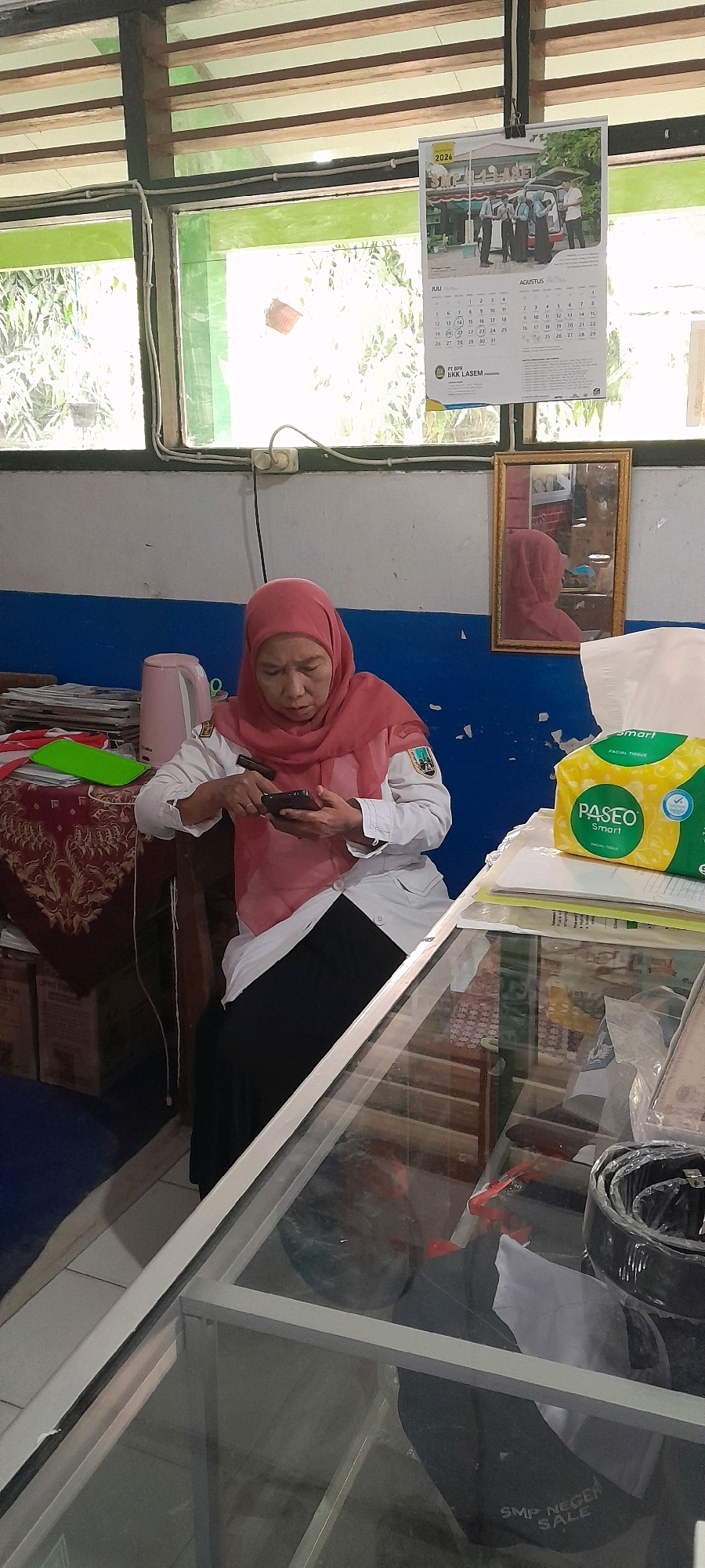A middle-aged woman wearing a pink hijab and a white uniform with a black skirt is seated at a desk, looking down at her smartphone. The desk is cluttered with papers, a pink electric kettle, and a box of facial tissues. Behind her, a calendar with images of students hangs on the wall, dated 2026. A framed mirror reflects the woman's image. In the foreground, a glass display case contains various items, including a belt. The scene appears to be indoors, possibly a classroom or an office, with sunlight streaming through a window overlooking lush greenery. The overall impression is one of a typical day in a school or administrative setting in Tahunan, Indonesia.