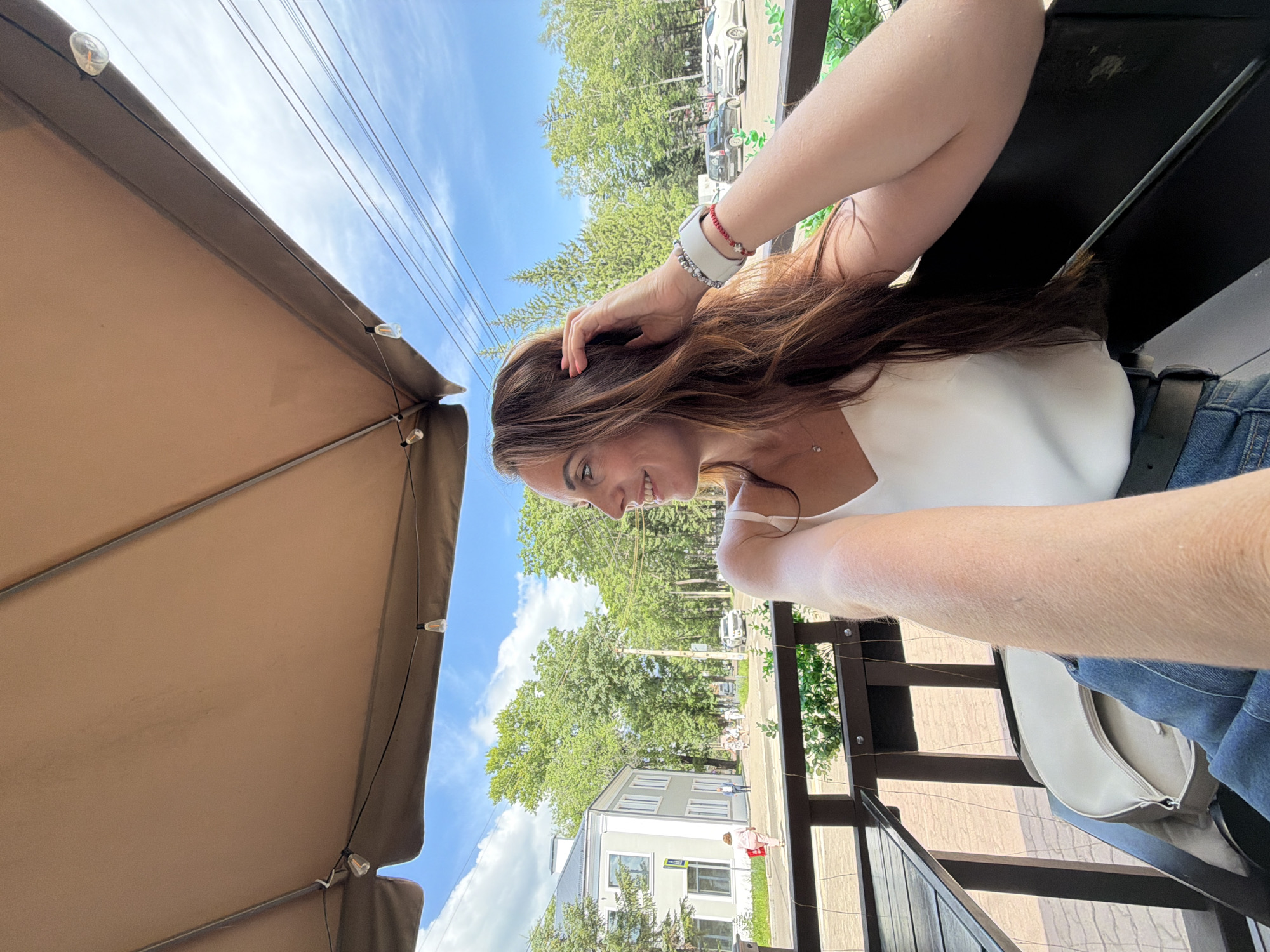 A woman with long brown hair is seated outdoors under a tan awning, looking to her left and smiling. She is wearing a white tank top and blue jeans. On her left wrist, she has a white smartwatch and a red bracelet. She is resting her right hand on her head. In the background, there is a street with a light-colored building, trees, parked cars, and utility lines against a blue sky with clouds. This scene is set in the city of Birobidzhan, Russia.