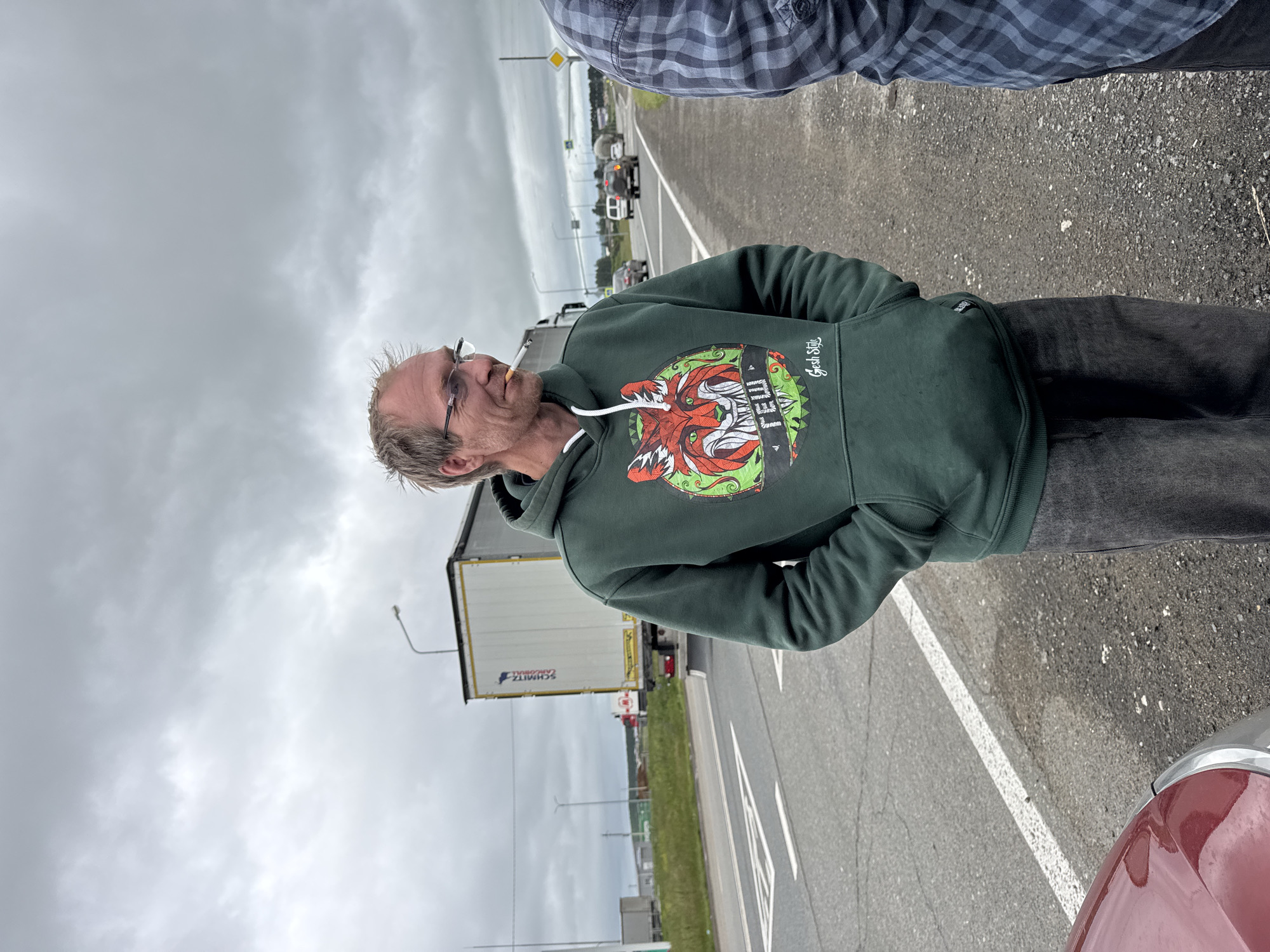 An overcast day at an exit or entrance to Alnashi, Russia, on a roadside. A Caucasian man with thinning blond hair, wearing glasses and a dark green hooded sweatshirt with a graphic of a red fox and the text "ГЕШ Gesh Style", stands with his hands in his pockets, smoking a cigarette. He is on the side of a road with multiple lanes, a white line marking the edge of the road, and an off-ramp visible in the distance. A large white semi-trailer truck, labeled "SCHMITZ CARGOBULL", is parked on the side of the road behind him. Several other vehicles are visible on the road, including cars and a cement mixer truck. The sky is cloudy and gray. Another person, wearing a blue and gray plaid shirt, is standing to the right of the frame, partially visible. A portion of a red car is visible in the lower left corner of the image.