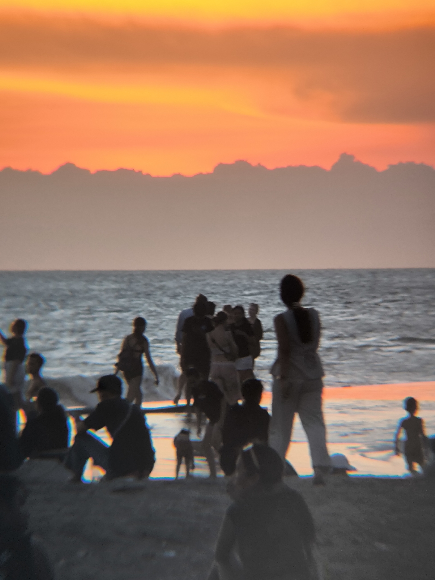 This image depicts a crowded beach scene in North Kuta, Indonesia during sunset. The sky is vibrant with orange and yellow hues, illuminating a line of dark silhouettes along the horizon. The sea reflects the sunset's colors. In the foreground, numerous people are visible, their figures blurred and indistinct. Some people appear to be standing in the water, while others are sitting or walking on the sand. One person stands out with their back facing the viewer, wearing light-colored clothing. A small dog is visible near the center. The scene is one of a relaxed, social gathering at sunset.