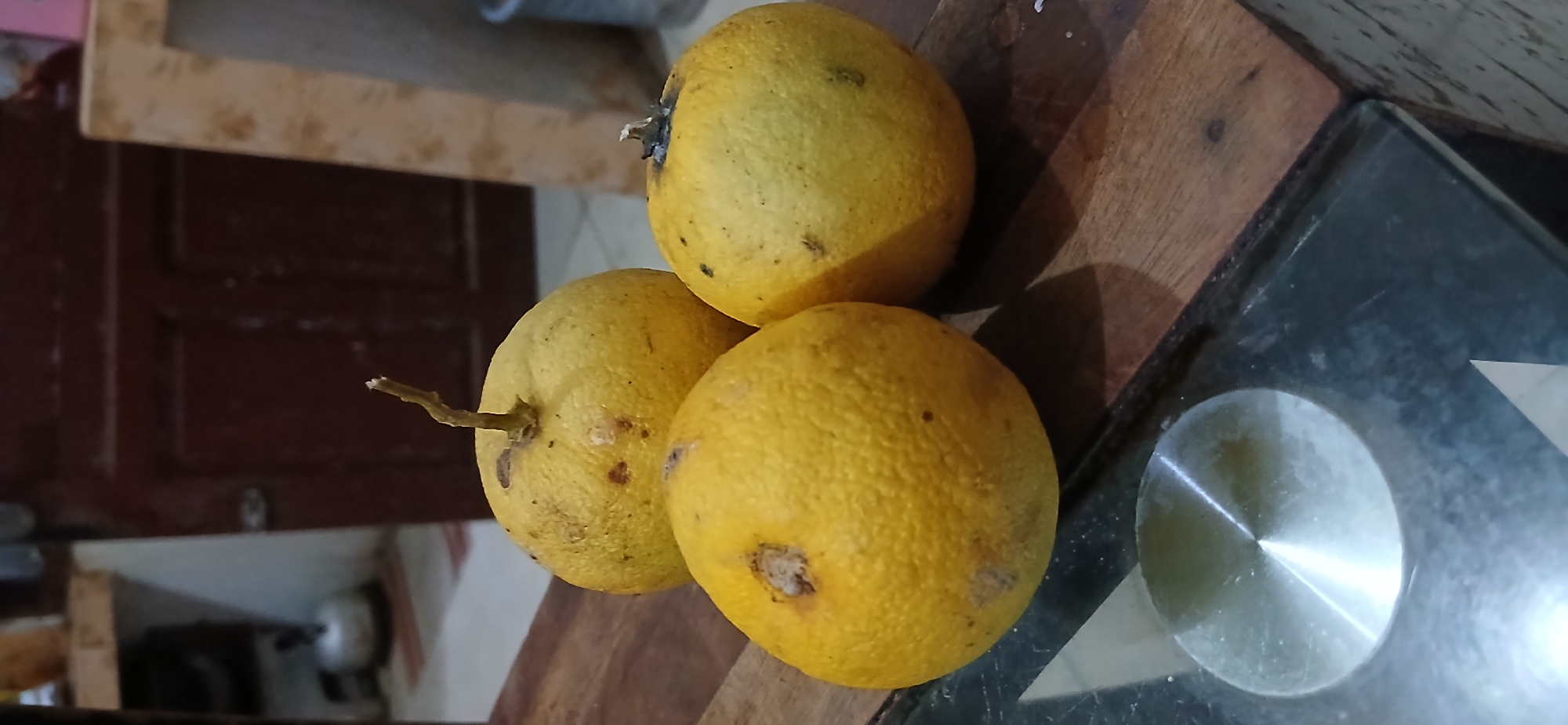 Three yellow, textured fruits, possibly oranges or lemons, are arranged on a wooden surface. The fruit in the foreground, on the left, is partially visible and closest to the camera. To its right and slightly behind, another fruit is more fully in view. The third fruit, positioned behind and to the right of the other two, is also fully visible. The wooden surface appears to be a table. The background shows a dark wooden door and some kitchen elements, including what looks like a kettle. The scene is indoors, likely in a domestic setting within Samarinda, Indonesia, as suggested by the context of the image. The lighting is dim, casting subtle shadows.