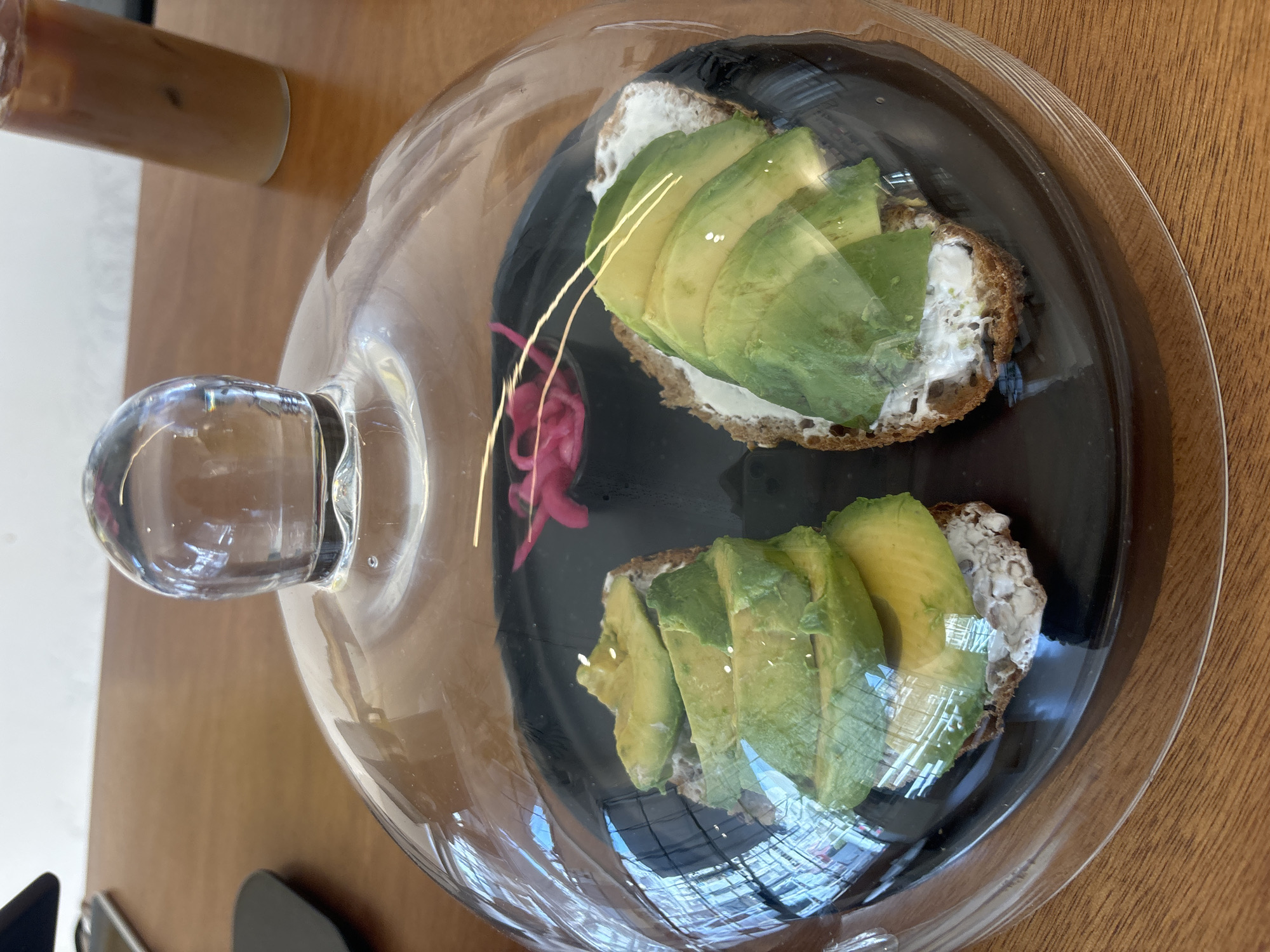 The image depicts two avocado toasts under a clear glass dome on a black plate. The toasts appear to be topped with a white spread and sliced avocado. There are some pink pickled onions nearby the toast at the top of the photo. A tall glass with a light brown drink is present on the left side of the frame. The surface beneath appears to be a wooden tabletop. While it is impossible to confirm the location without additional context, there are no definitive visual cues within the image to determine whether it is in Tel-Aviv, Israel.