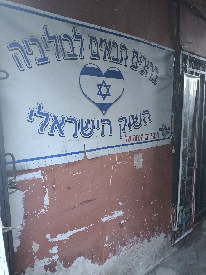 A sign in Hebrew reads "Welcome to Bolivia" above a heart containing the Star of David, an image of the Israeli flag. Below this is "The Israeli Market". The sign is affixed to a wall with peeling brown paint. To the right of the sign, a barred window or door is visible. The overall scene appears weathered and somewhat run-down.
