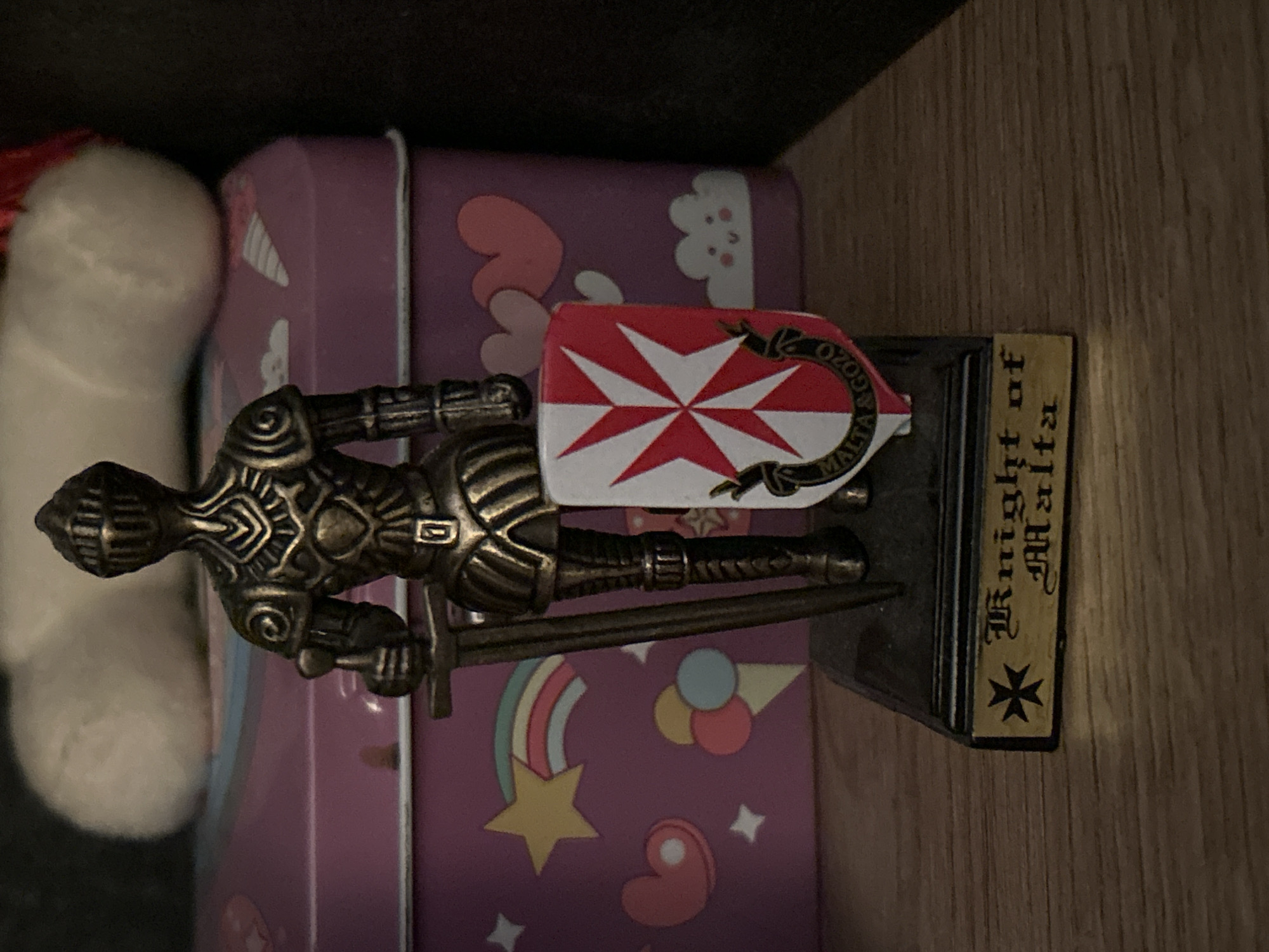 A close-up, eye-level, full-shot photograph shows a statue of a knight holding a sword and shield. The statue is made of dark metal and stands on a black base with a gold plaque that reads "Knight of Malta." The shield has the Cross of Malta design, with the words "Malta & Gozo" around the bottom. The statue is positioned on a wooden shelf in front of a purple, decorative tin box. The tin box is adorned with colorful drawings of rainbows, stars, hearts, and ice cream cones. A blurred, white, fluffy object is visible behind the tin box. The lighting is dim, casting shadows and highlighting the metallic texture of the statue. The scene appears to be indoors, possibly a display shelf in a home or shop. There are no people present in the image, and no specific location in Sofia, Bulgaria is indicated.