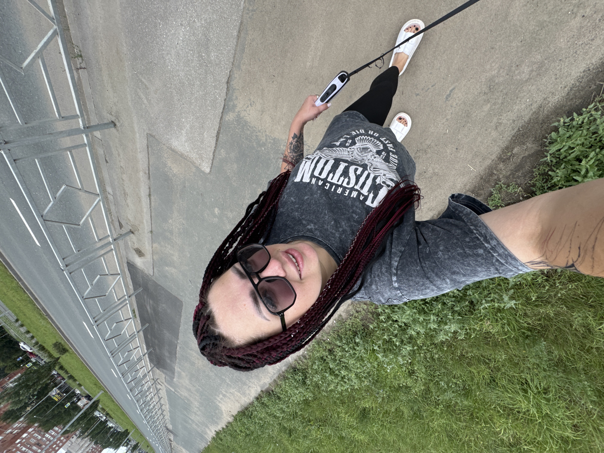 In Tolyatti, Russia, on a cloudy day, a woman with dark braided hair and sunglasses is captured in a high-angle selfie walking on a sidewalk. She is wearing a gray t-shirt with a graphic print, black leggings, and white slide sandals. In her right hand, she holds a black and white retractable dog leash. To her left, a patch of green grass runs alongside the sidewalk, which is bordered by a metal fence. Beyond the fence, a paved road with a white lane marking is visible, with cars and a multi-story building in the background.