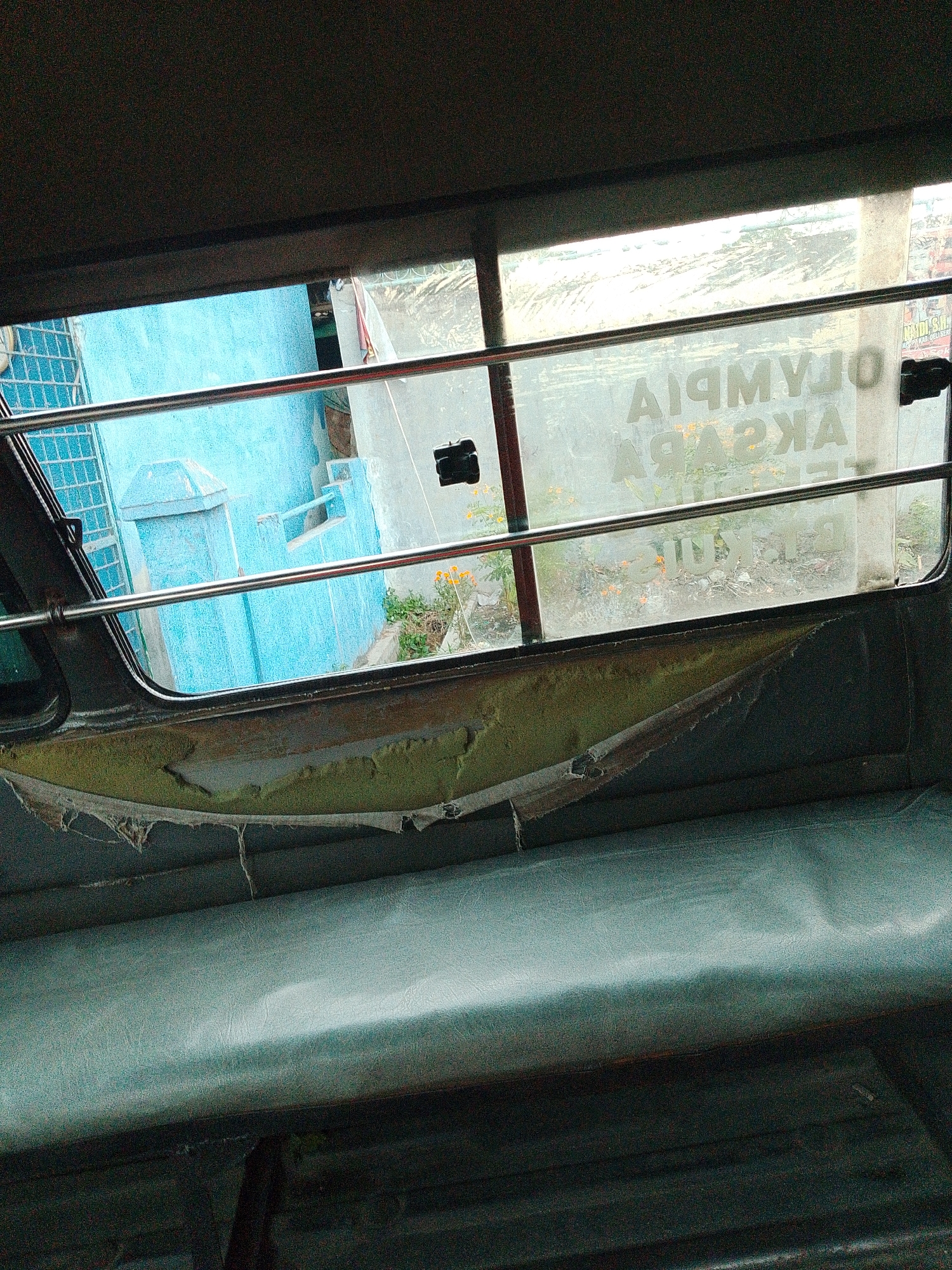 The image captures an interior view from what appears to be a public transportation vehicle, possibly a jeepney or bus, looking out of a window. The window is divided by a metal bar, and the glass itself is dirty and somewhat opaque, with some lettering visible on the exterior. Outside the window, a blue building with geometric patterns and architectural details is prominent. To the right, there is a glimpse of other structures and vegetation, including some orange flowers. The interior of the vehicle shows a worn-out seat made of a light blue, possibly vinyl material. The padding beneath the window appears damaged and is peeling away. The lighting suggests it is daytime. The text on the window is backwards and reads "OLYMPIA" and "AKAZPA", likely advertisements or signs from the exterior. The scene suggests an urban or semi-urban environment in Bandar Khalipah, Indonesia, characterized by modest infrastructure and signs of wear and tear.