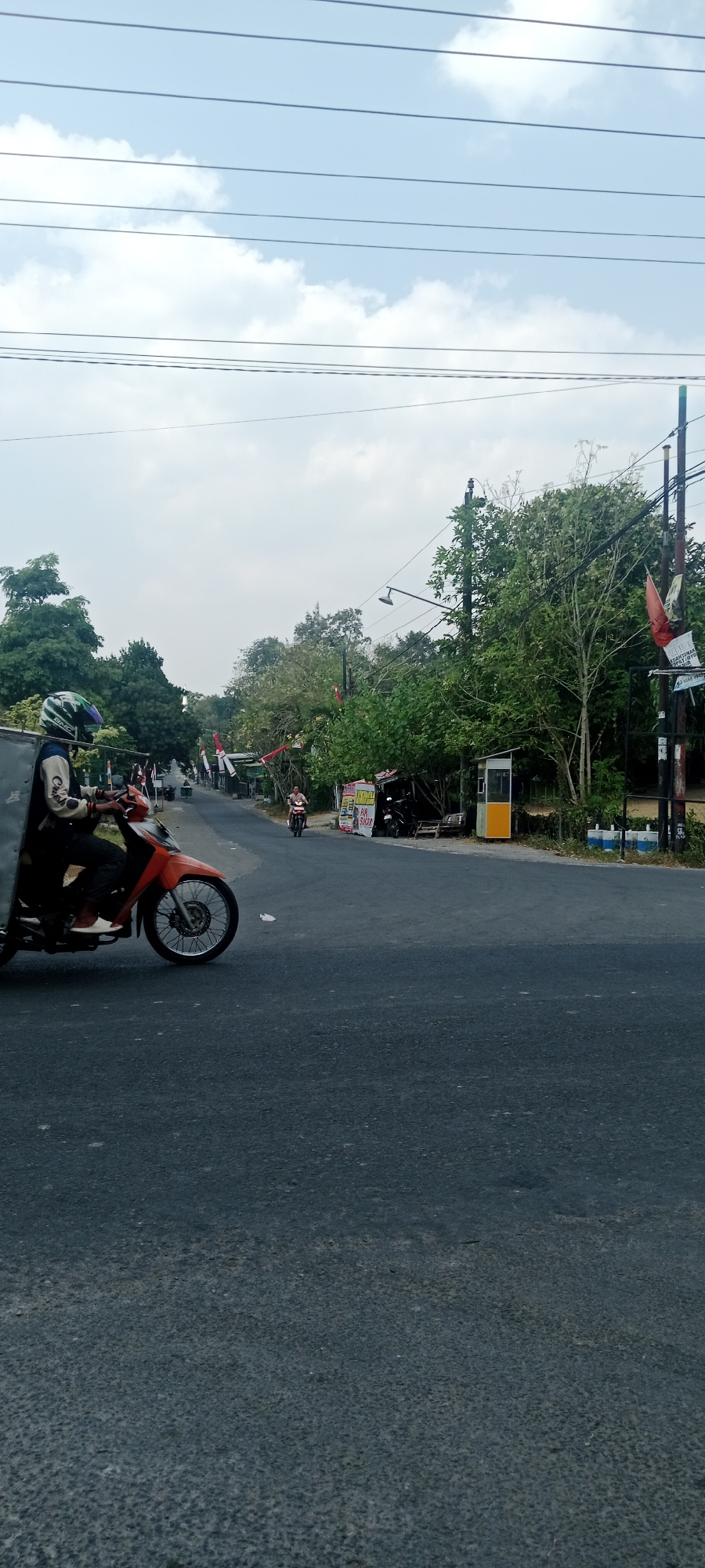 A person wearing a helmet rides an orange motorcycle on a paved road. The person is wearing a jacket and pants. The motorcycle has a box attached to the back. The road is bordered by trees and a few buildings. There are some red and white flags visible along the side of the road. Overhead, power lines stretch across the sky, which is partially cloudy. The scene appears to be in Maguwoharjo, Indonesia.