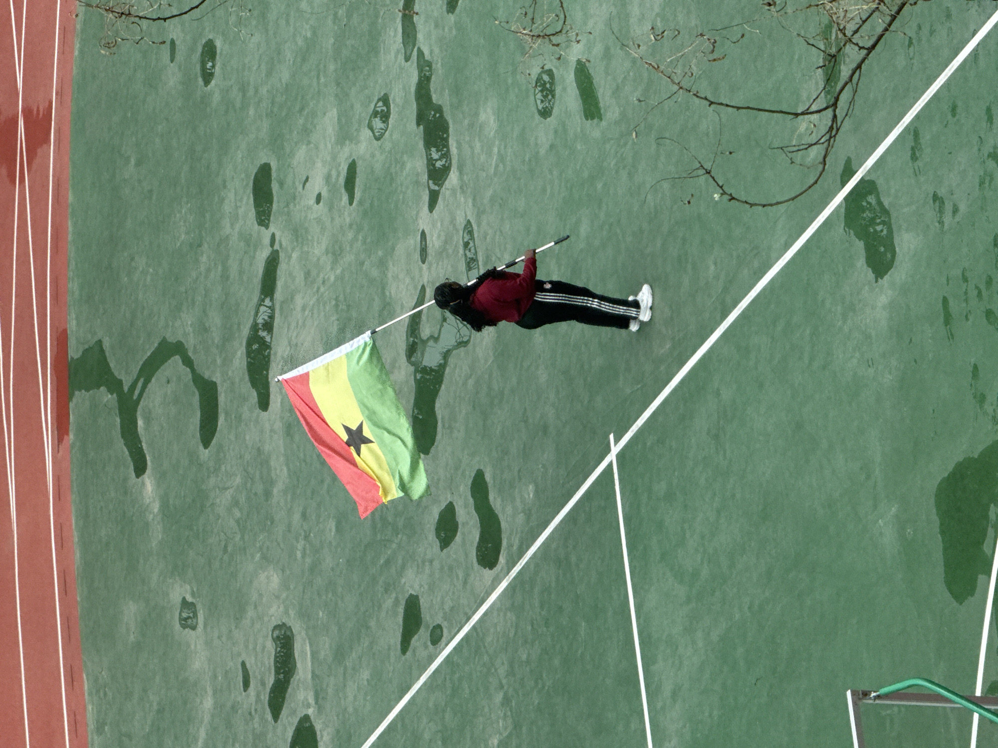 An aerial, high-angle shot captures a person lying on their back on a wet, green sports court, holding up a Ghanaian flag on a pole. The scene is an outdoor sports facility, likely in Daxing District, China.
The lone individual, possibly female due to long dark hair, is dressed in casual athletic wear: a dark red long-sleeved top, black track pants with white stripes down the sides, and white athletic shoes. They are positioned centrally on the court, holding the flag pole aloft with both hands. The Ghanaian flag, distinct with its red, gold, and green horizontal stripes and a prominent black star in the center, unfurls above the person.
The court surface is a muted green, appearing damp with numerous dark, irregularly shaped patches and streaks that suggest water puddles or recently wet areas, indicating that it has recently rained or is currently drizzling. To the left of the frame, a segment of a red running track with white lane markings is visible. In the upper part of the image, the bare, leafless branches of a tree extend into the frame, suggesting an autumn or winter season. White boundary lines crisscross the green court, delineating different play areas. A green structure, possibly part of a goalpost or net, can be seen at the bottom right corner. The lighting is soft and even, characteristic of an overcast day. There is no visible text other than the flag's design.