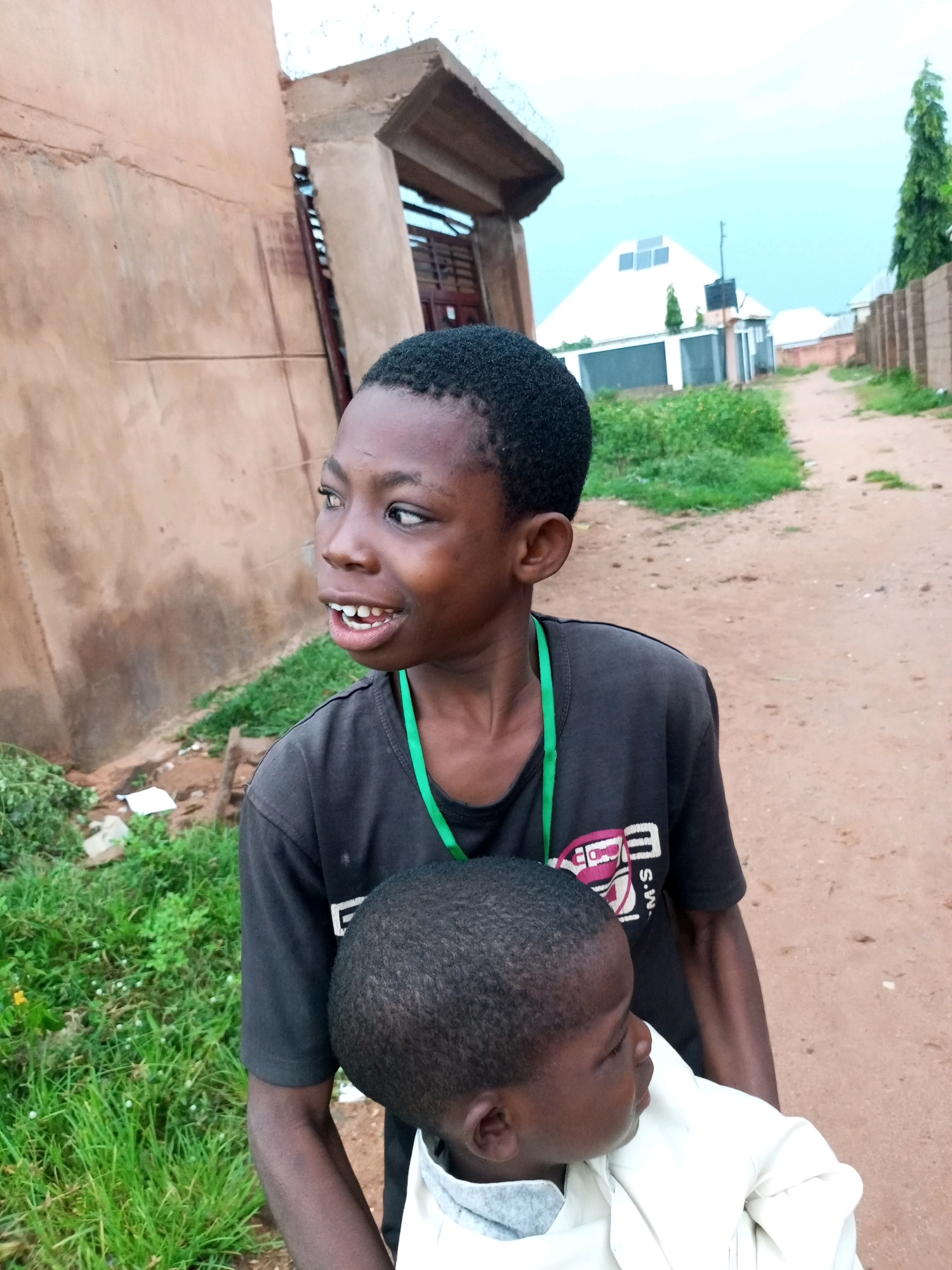 In the city of Gombe, Nigeria, two young boys are captured in a photograph outdoors. The elder boy, positioned in the foreground, has dark skin and short, black hair. He is wearing a dark t-shirt with a green lanyard around his neck. His mouth is slightly open, revealing his teeth, and he is looking to his left. He appears to be holding a younger child, whose head and upper body are visible, wrapped in a light-colored garment. The background shows a dirt road and a building with a rough, textured wall. Further in the distance, there are more structures and lush green vegetation, suggesting a residential or semi-urban environment. The sky is overcast with a pale blue hue.