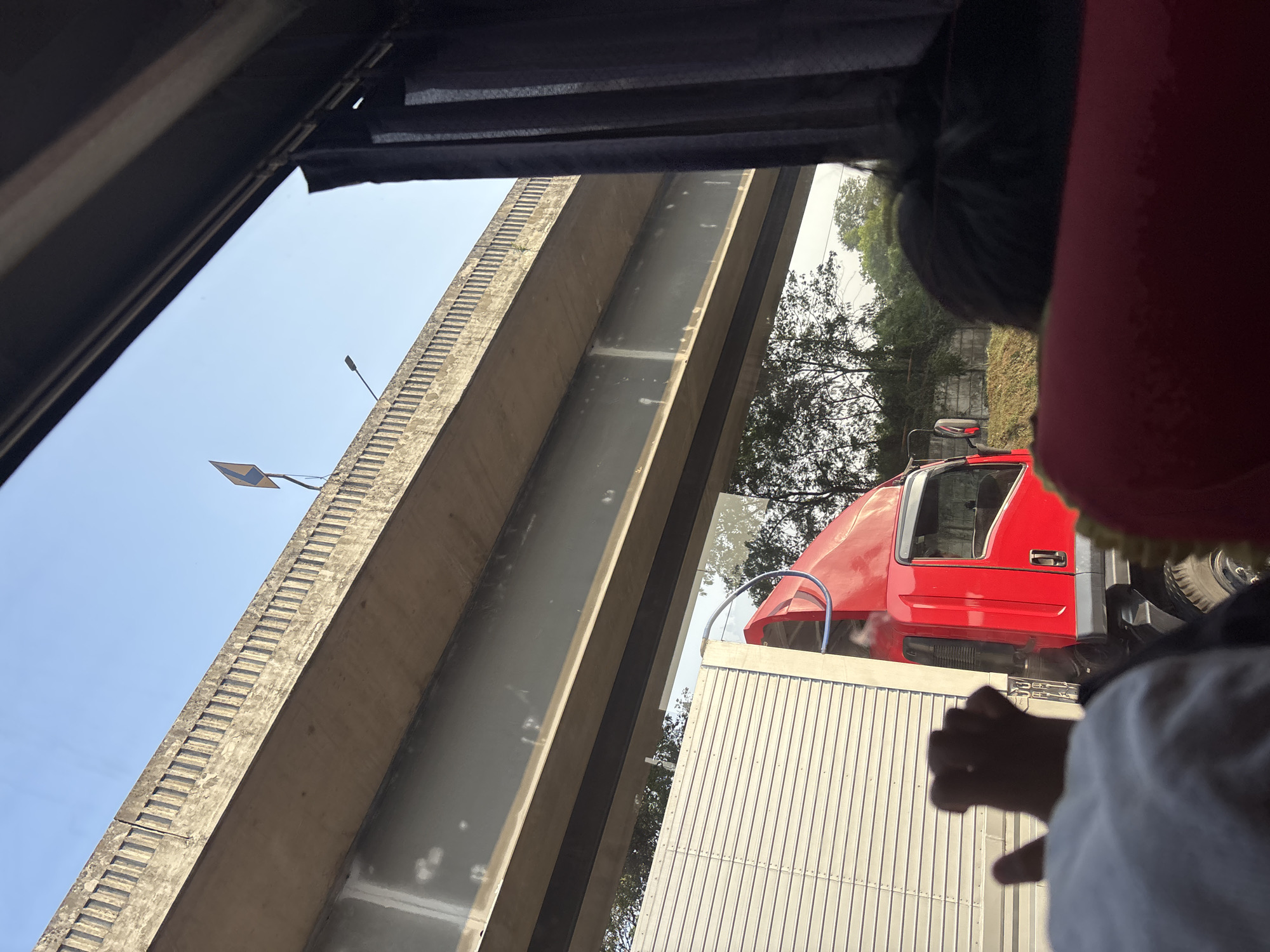 An overpass runs diagonally across the frame from the upper left to the lower right. Underneath the overpass, a red truck with a white trailer is stopped, partially visible on the right side of the image. The truck's cabin is bright red, and its trailer is made of corrugated metal. The scene appears to be on a clear day, with a pale blue sky visible above the overpass. In the foreground, the back of a person's head is visible, and a hand is reaching out to the left. The image is taken from inside a vehicle, with dark curtains visible on the right side of the frame, suggesting the viewer is a passenger. The presence of an overpass and a large truck suggests an urban or industrial area. The specific location in Karawang, Indonesia, is suggested by the context of the request, but not visually confirmed by the image itself.