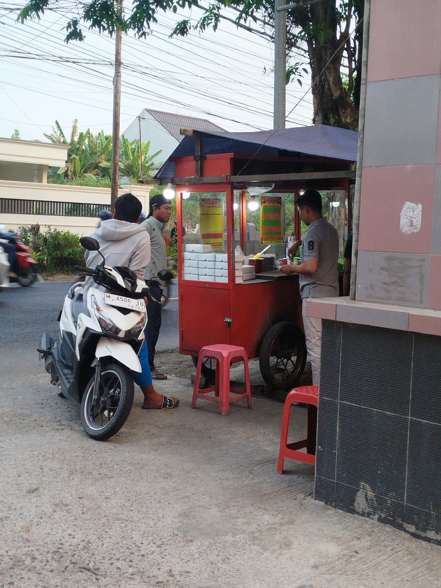 In Burneh, Indonesia, a street food stall vendor, dressed in a gray polo shirt and white pants, is serving a customer. The vendor is positioned behind a red food cart under a purple awning, with bright lights illuminating the area. He is in the process of handling something over the counter. Beside the cart, a white motorcycle is parked. A young man wearing a gray hoodie and blue shorts sits on the motorcycle, looking towards the vendor. Another man, wearing a black cap and a gray shirt, stands near the vendor, observing. Two red plastic stools are placed near the stall. The scene suggests a casual interaction at a street-side eatery. The time appears to be late afternoon or early evening, with the sky a muted gray and soft lighting. The background shows a residential building with a fence and lush green banana trees, indicating a tropical environment. The motorcycle's license plate, M 4506 JD, is visible.