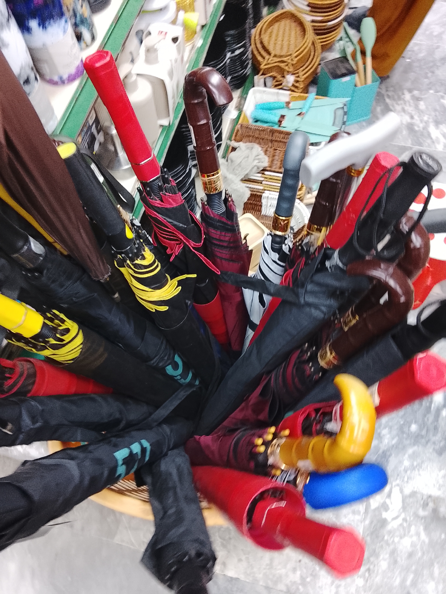 The image shows a collection of umbrellas with various handle styles (straight, curved) and colors (red, black, brown, yellow) in a container, possibly a basket, inside what appears to be a store in Gulgasht Colony, Pakistan. Some umbrellas are closed, with the fabric cover visible. The environment includes other merchandise such as baskets, kitchen utensils, and other household items. The image does not include any people or interaction. The focus is on the collection of umbrellas.