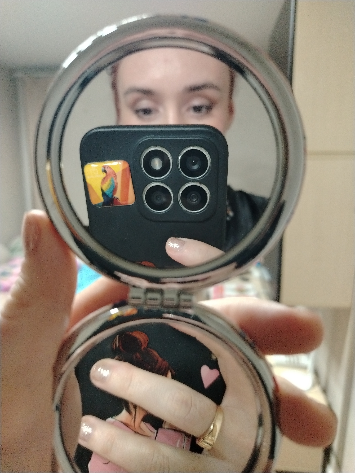 A person is holding a round, hinged compact mirror, reflecting their face and the smartphone they are using to take the picture. The smartphone has a black case with a sticker of a parrot on the back. The camera of the phone has four lenses. The person's face is visible in the top mirror of the compact, and their fingers, with a ring on one, are reflected in the bottom mirror of the compact, showing an image of a person with their hair in a bun and a pink heart shape. The fingernails are painted with a shimmery polish. The background is blurry, but a wall, possibly in a room, is visible. There is no information to determine if this image was taken in Izhevsk, Russia.