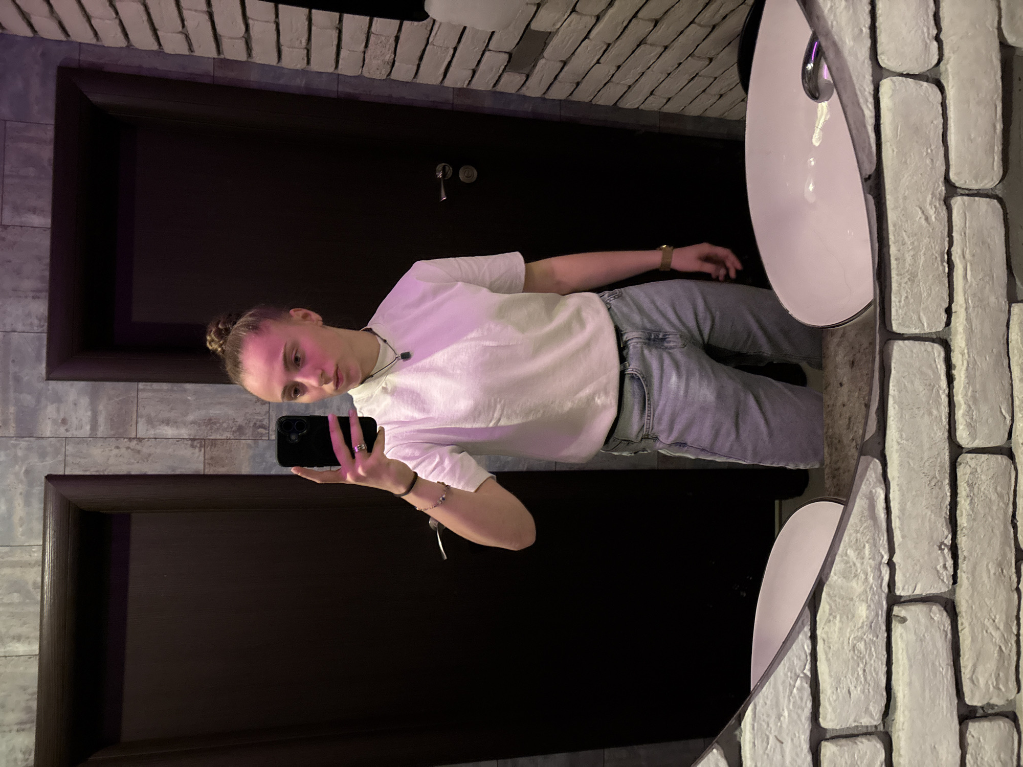 A person is taking a selfie in a bathroom with a brick wall on the right and a dark brown door with a frame on the left. The person is wearing a white t-shirt and light blue jeans. There is a sink in the corner. The exact location in Sofia, Bulgaria cannot be determined from the image.