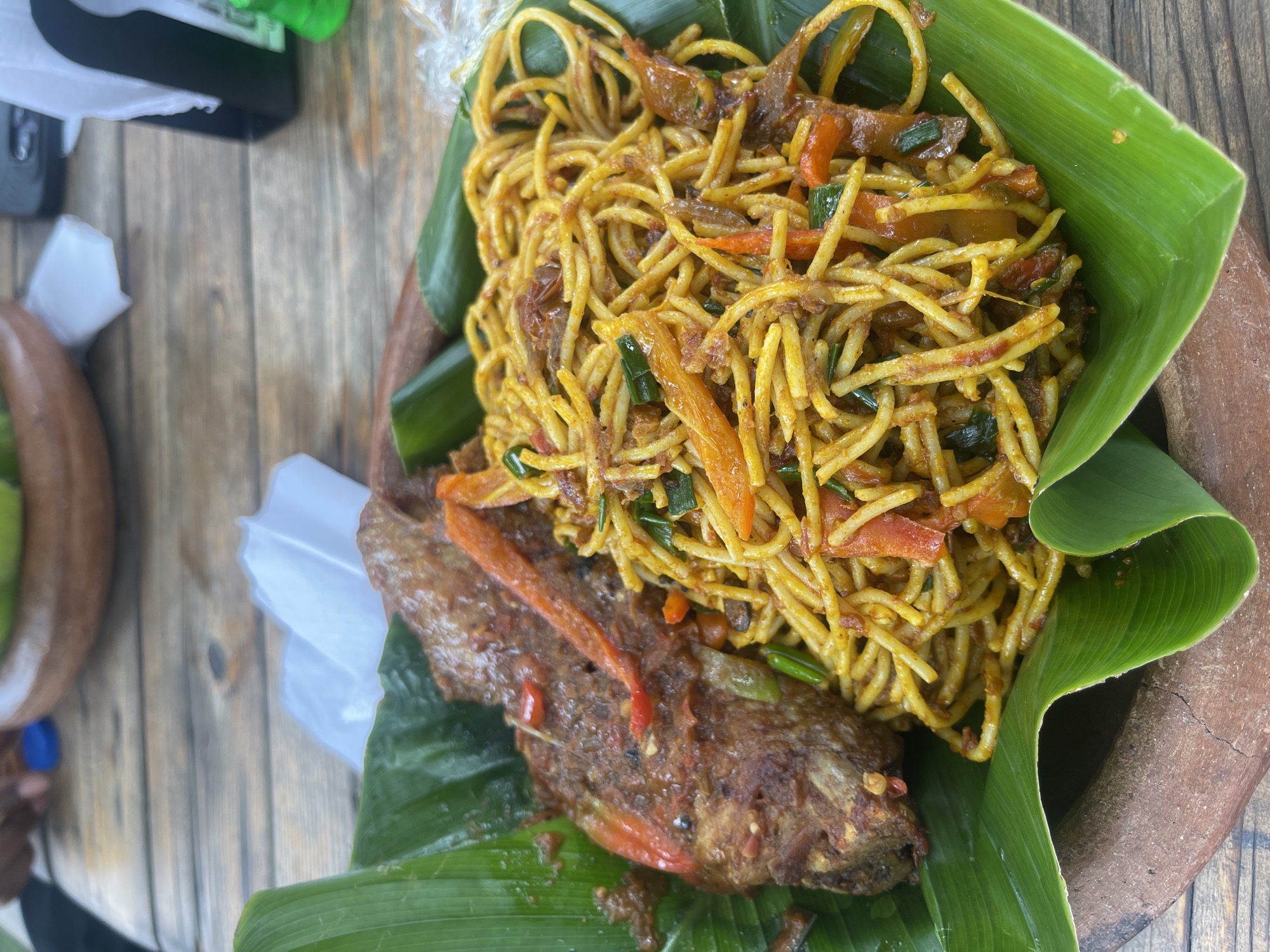 A top-down shot shows a plate of food, possibly in Abuja, Nigeria, based on the cultural presentation. The food is served on a large green banana leaf, placed in a shallow brown bowl. The meal consists of fried spaghetti with vegetables and a fried fish. The spaghetti is yellow-orange and mixed with strips of carrots, green onions, and bits of fried onions or meat. The fish is reddish-brown, possibly seasoned with spices, with visible red pepper strips. A white plastic bag is present to the left of the fish. The wooden table is visible underneath.