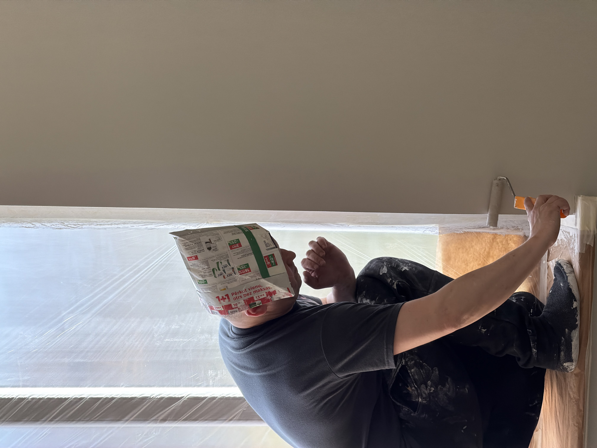 A man is painting an interior wall. He is wearing black pants and a gray shirt, both covered in paint splatters. He has a newspaper or advertisement page covering his head. He is using a paint roller with an orange handle to apply paint to the wall. The window is covered with plastic film. The OCR text on the man's head is in Latvian, with product sale information. The location appears to be an indoor residential room, possibly in the city of Dreiliņi, Latvia based on the OCR language.