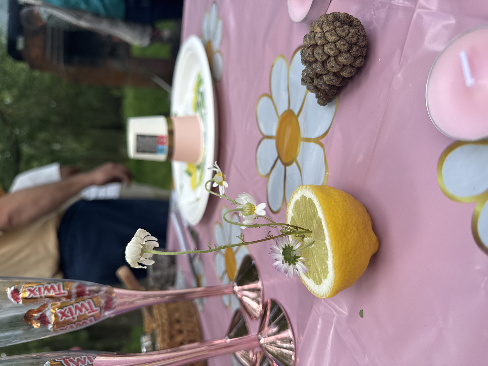 The image shows a pink tablecloth with a daisy pattern. On the table are various objects including a halved lemon with small flowers inserted, a pine cone, a pink candle, a white plate with a pink container on top, and champagne flutes filled with Twix candy bars. Part of a person is visible on the left side of the image, seated at the table. The background shows greenery, suggesting an outdoor setting. There is no explicit information to indicate this image was taken in Burgas, Bulgaria.