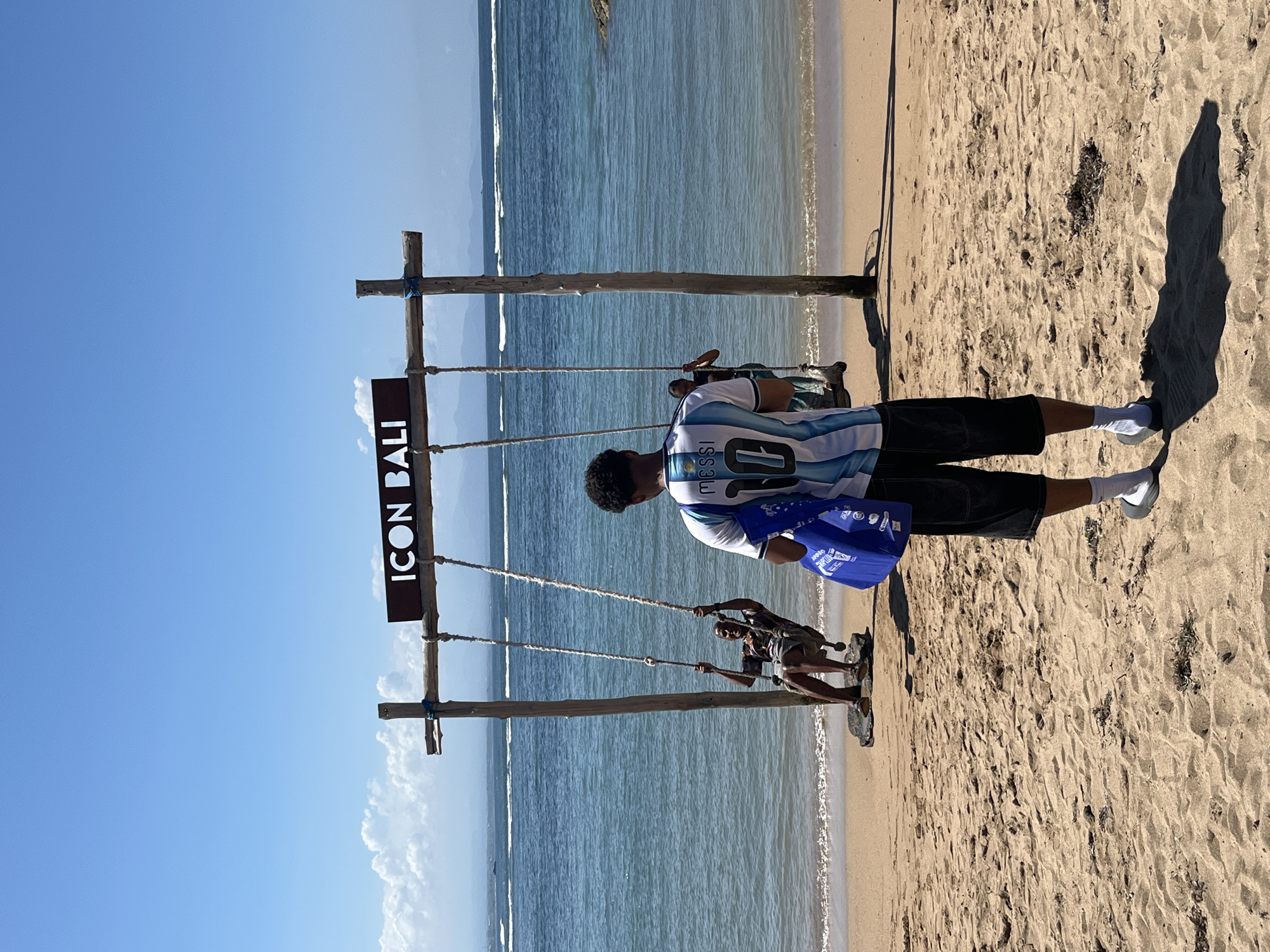 A man stands on a sandy beach in front of the ocean, facing away from the camera. He is wearing a white and blue striped jersey with the name "MESSI" and the number "10" on the back, along with black shorts, white socks, and grey sandals. He is holding a blue bag in his left hand. In the background, two people are on swings that are suspended from a wooden frame. The frame has a sign that reads "ICON BALI". The ocean is calm with small waves and a clear blue sky overhead. The location is likely on a beach in Denpasar, Indonesia, given the sign.