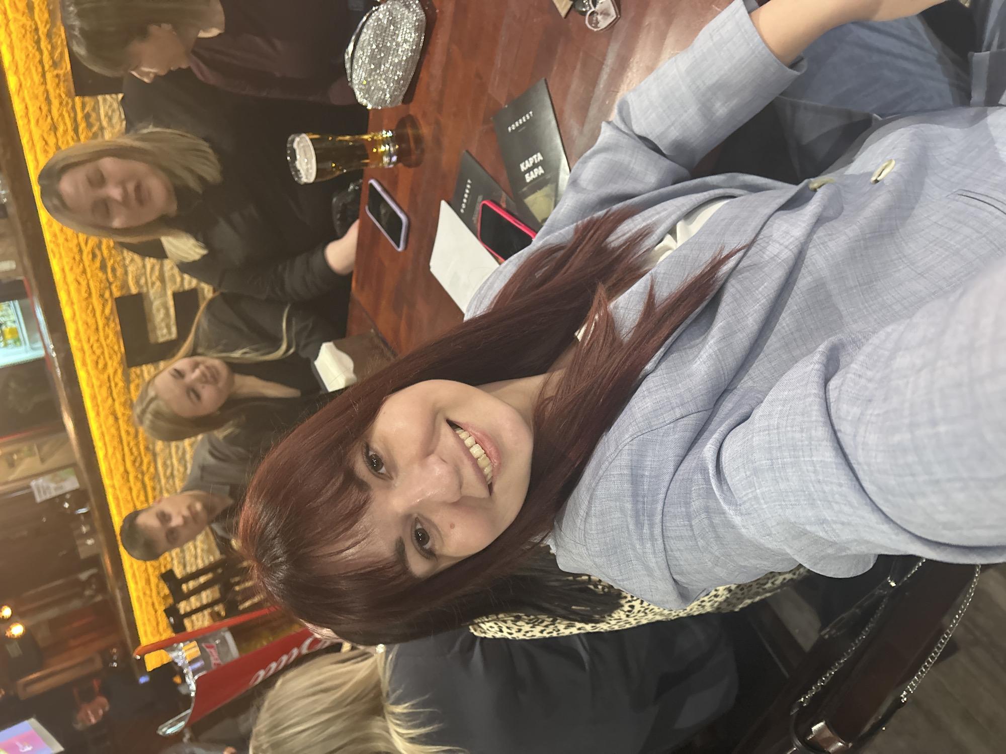 The image shows a group of people at a table in what appears to be a bar or restaurant. A woman with red hair and a light blue blazer is in the foreground, seemingly taking a selfie or posing for the camera. In the background, there are other people, drinks (possibly beer), a menu with text in Russian, and phones. The menu reads "FORREST KARTA BAPA" which is likely related to the Forrest Restaurant.
Based on the menu and the information provided, the location is the city of Chaikovsky, Russia. The atmosphere is casual, suggesting a social gathering or outing.