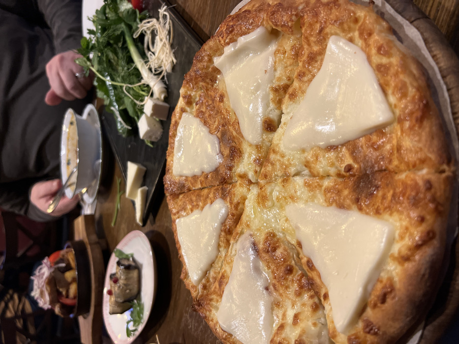The image displays a dining scene inside a Georgian restaurant in Saint Petersburg, Russia. The foreground is dominated by a large, round Imeruli-style khachapuri, baked golden brown with six triangular slices of melted white cheese on top, resting on a rustic wooden board. The khachapuri is pre-cut into eight radial slices.
On the wooden table, further back and to the left, a small white plate with a pink rim holds two eggplant rolls (likely Badrijani Nigvzit) garnished with a green sprig and small red berries. Behind it, a small cast-iron pan on a wooden stand contains a dish of roasted potatoes or meat and potatoes, topped with thinly sliced red onions.
To the right of the eggplant dish, a black rectangular plate features an assortment of fresh herbs including dill, parsley, and green onions, accompanied by various white cheeses – some cut into small cubes and others into flat slices.
In the mid-background, two blurred sets of hands are visible. One hand, wearing a ring on the fourth finger, holds a spoon in a white bowl containing a light-colored soup, which rests on a saucer. The individual is wearing a gray long-sleeved shirt. Another person's arm and shoulder are partially visible on the far left. The table surface is a polished dark wood, and the lighting is subdued, suggesting an indoor, evening dining atmosphere.