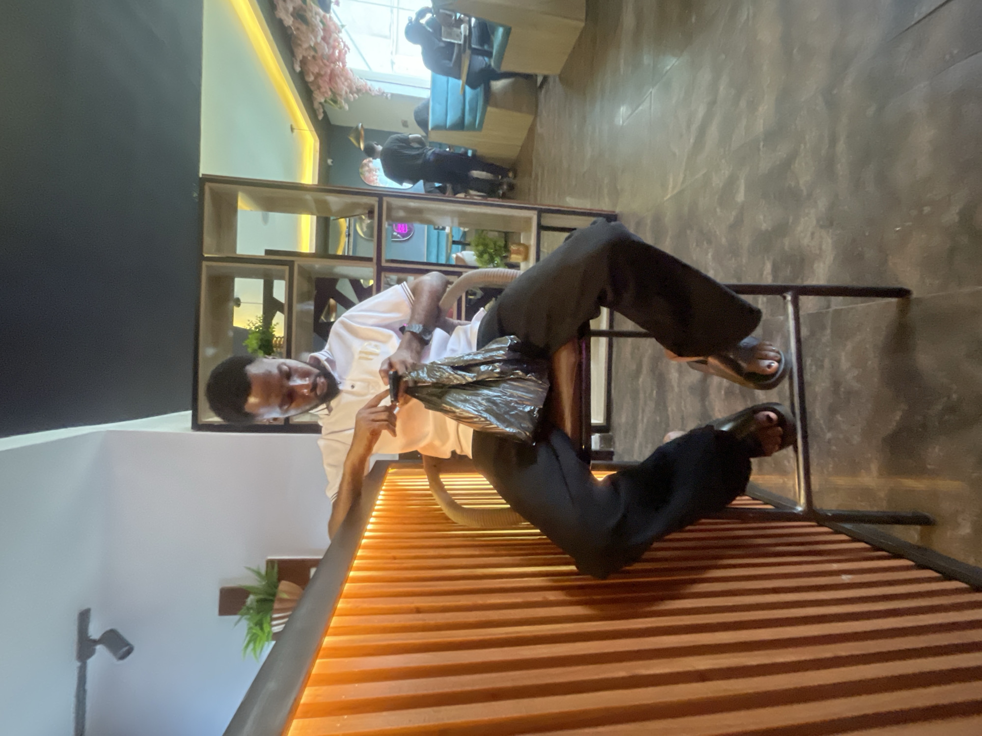 A medium shot captures a man seated on a metal-framed chair in what appears to be a commercial establishment, possibly a cafe or restaurant, located in Adankolo, Nigeria. The man, dressed in a white, short-sleeved collared shirt and black pants, is focused on an object wrapped in a shiny, dark material, possibly plastic or metallic, which he holds in his hands. He is fair-skinned, has short dark hair, and is wearing a watch on his left wrist. The man's feet are clad in sandals.
The establishment is furnished with booth seating and a shelving unit in the background. Other patrons are visible in the background. A decorative plant is mounted on the wall behind him. The flooring is a textured grey material. The man's chair is positioned near a wooden slat barrier, likely for dividing space.