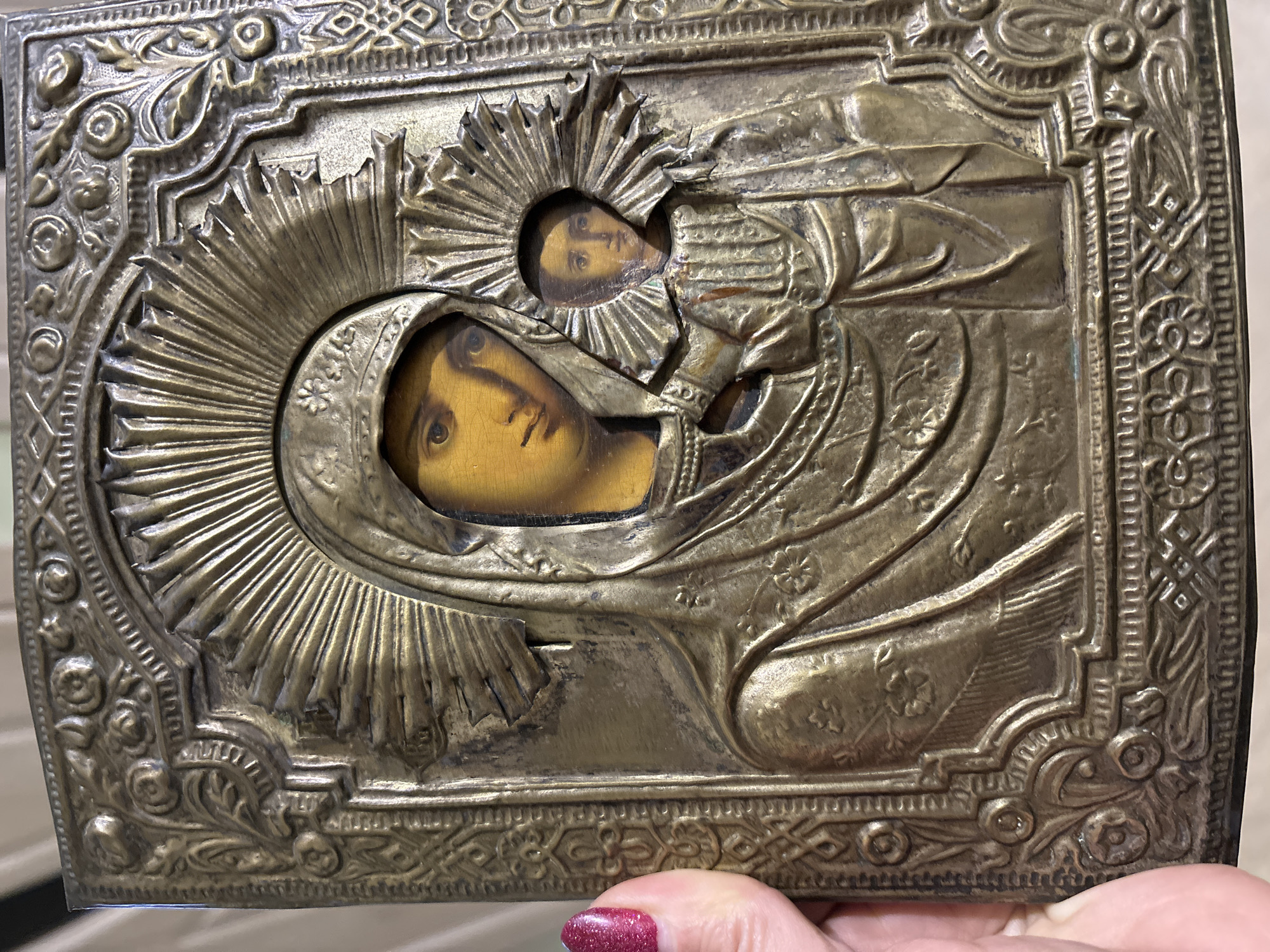 An antique Russian Orthodox icon, depicting the Theotokos (Virgin Mary) and Christ Child, is presented in a close-up, angled view. The icon features a painted wooden panel covered by an ornate, tarnished brass or bronze oklad (metal riza) that leaves the faces and hands of the figures exposed. The Virgin Mary, depicted on the left, wears a veiled headdress, her face visible with a gentle expression and eyes gazing slightly to the right. To her right, the Christ Child is shown, his face looking directly forward, with his right hand raised in a blessing gesture. Both figures are surrounded by rayed halos on the oklad, with the Christ Child's halo exhibiting damage and distortion to the metal rays. The oklad features intricate embossed patterns, including floral motifs and geometric designs along its borders. The metal surface shows significant patina, discoloration, and wear consistent with age. A human hand, with a polished, sparkling pink fingernail, holds the icon by its left edge, indicating it is being examined or displayed. The background is out of focus, suggesting an indoor setting with a light-colored, possibly textured surface. The scene takes place in Chaikovsky, Russia.