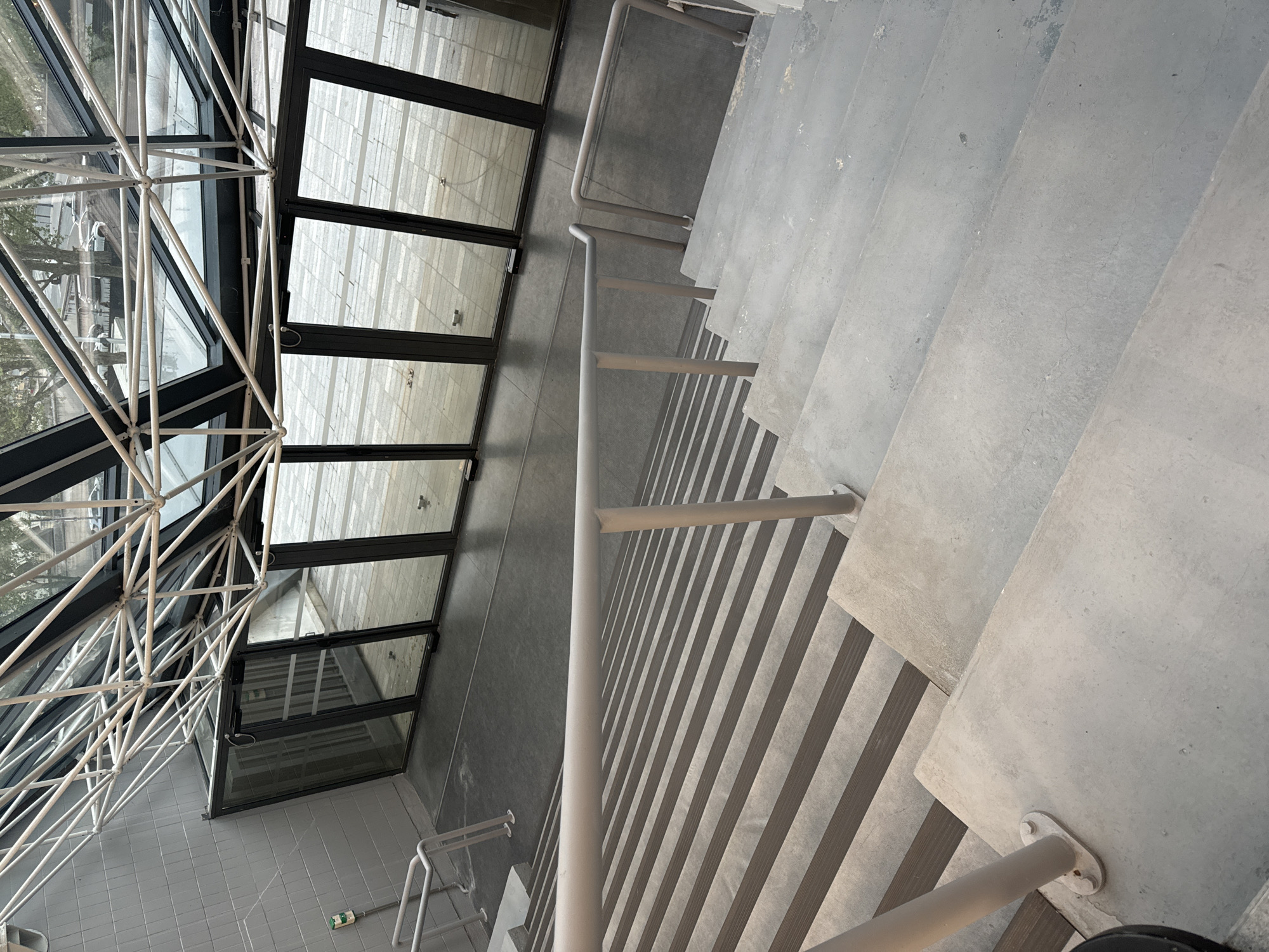 The image depicts a modern interior space with a combination of architectural features. A glass ceiling or skylight structure, supported by a white metal framework, dominates the left side of the frame. Adjacent to this is a series of tall, vertical windows or glass doors with black frames.
To the right, a concrete staircase descends out of view. A white handrail runs alongside the stairs. The walls and flooring appear to be finished in a light gray or off-white concrete. There are no visible people in the scene.
The scene is modern and appears to be taken indoors. Based on the architectural style and the presence of the glass ceiling, the location could be in Paris, France. The glass ceiling is reminiscent of contemporary architectural designs often found in public or commercial spaces in Paris.