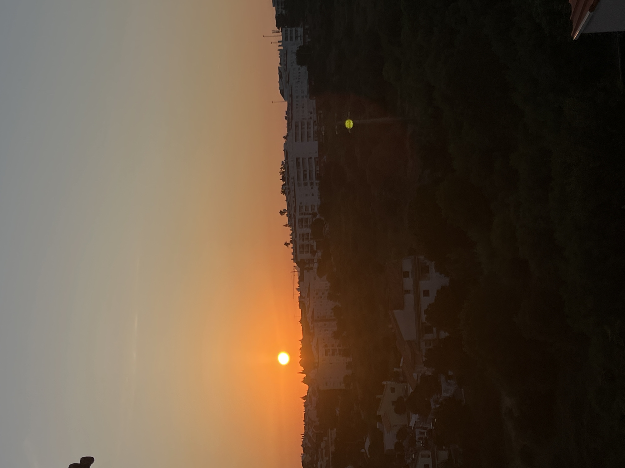 A sunset is depicted over the city of Cascais, Portugal. The sun, a bright orange orb, is positioned low in the sky, casting a warm glow over the scene. Silhouettes of buildings and trees are visible against the vibrant sky, suggesting an urban or suburban landscape. The foreground is dominated by dark shadows, obscuring details of the terrain and any potential inhabitants. The overall impression is one of a peaceful evening in a coastal city.