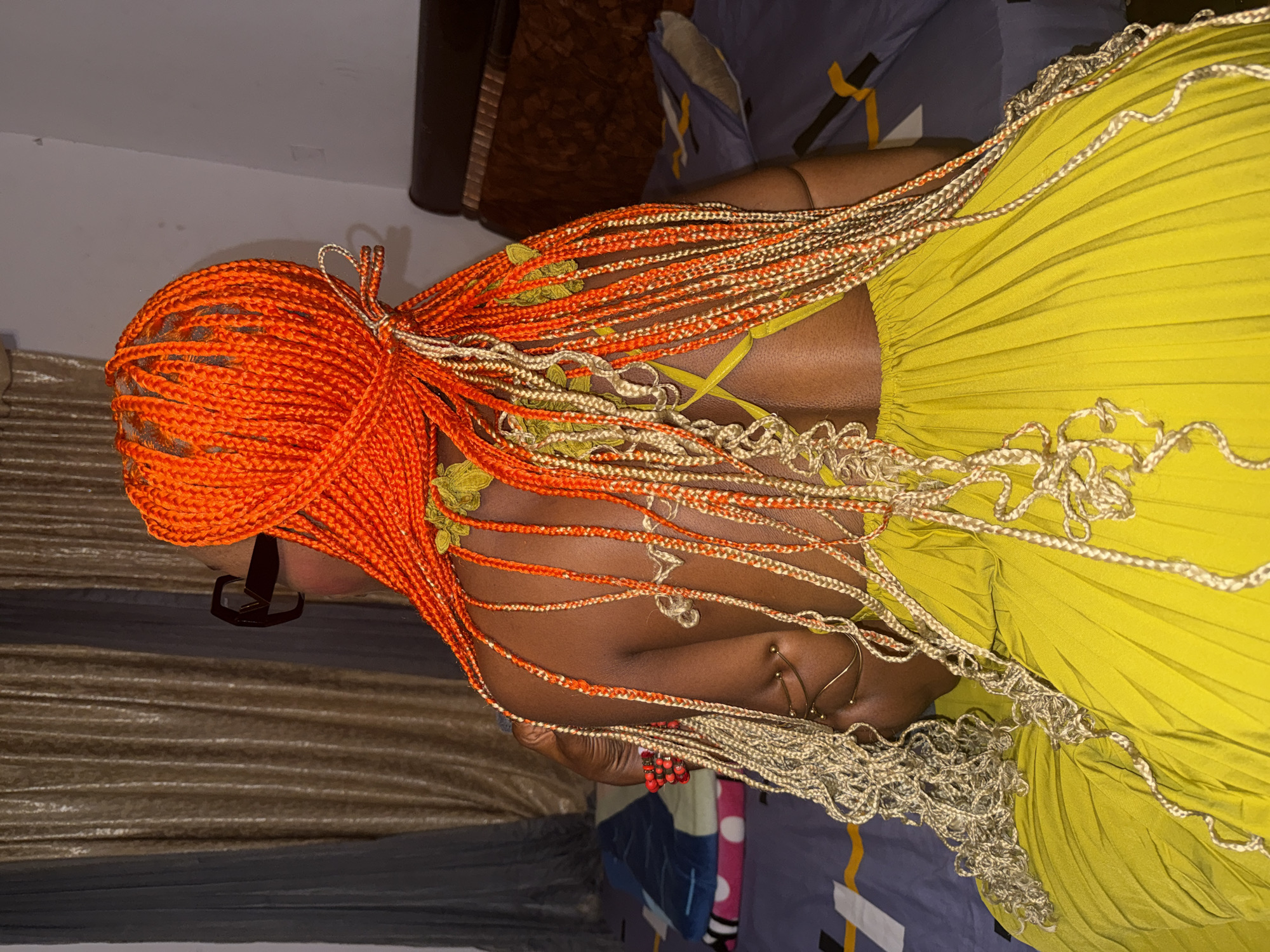 A person with bright orange braided hair, some strands of which are light blonde, is seen from the back, sitting on a bed in a room in Ado, Nigeria. They are wearing a bright yellow pleated dress. The person is also wearing dark-rimmed glasses. The bed has a dark brown headboard and is covered with a dark blue patterned duvet and a pink blanket. In the background, there are gold-colored curtains. The lighting suggests it is indoors, possibly at night or in a dimly lit room.