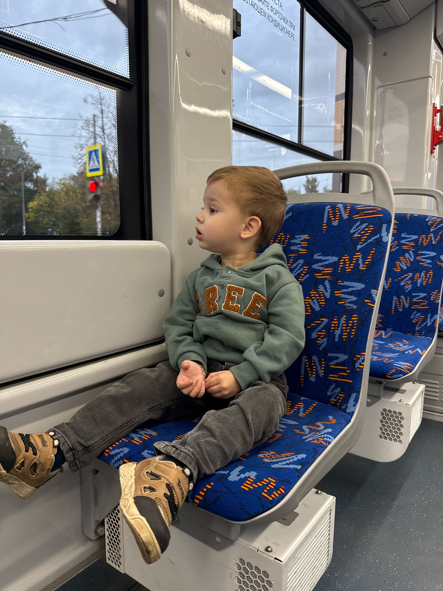 A young boy, appearing to be around 3-4 years old, sits on a blue and orange patterned seat inside a modern public transportation vehicle. He is wearing a teal hooded sweatshirt with "FREE" written in orange across the chest and dark grey pants. His feet, clad in brown and black athletic shoes, are crossed and resting on a lower platform. The boy is looking out of a window to his left, which shows a cloudy sky and some trees. A yellow pedestrian crossing sign and a red traffic light are visible through the window. The interior of the vehicle is clean and well-lit, with white paneling and grey seat frames. The seats are arranged in rows, and another identical seat is visible to the right of the boy, empty. The floor is a dark grey speckled material. The overall impression is a mundane scene of a child on public transport. The image does not contain any information that would allow for its location in Rusanovo, Russia.