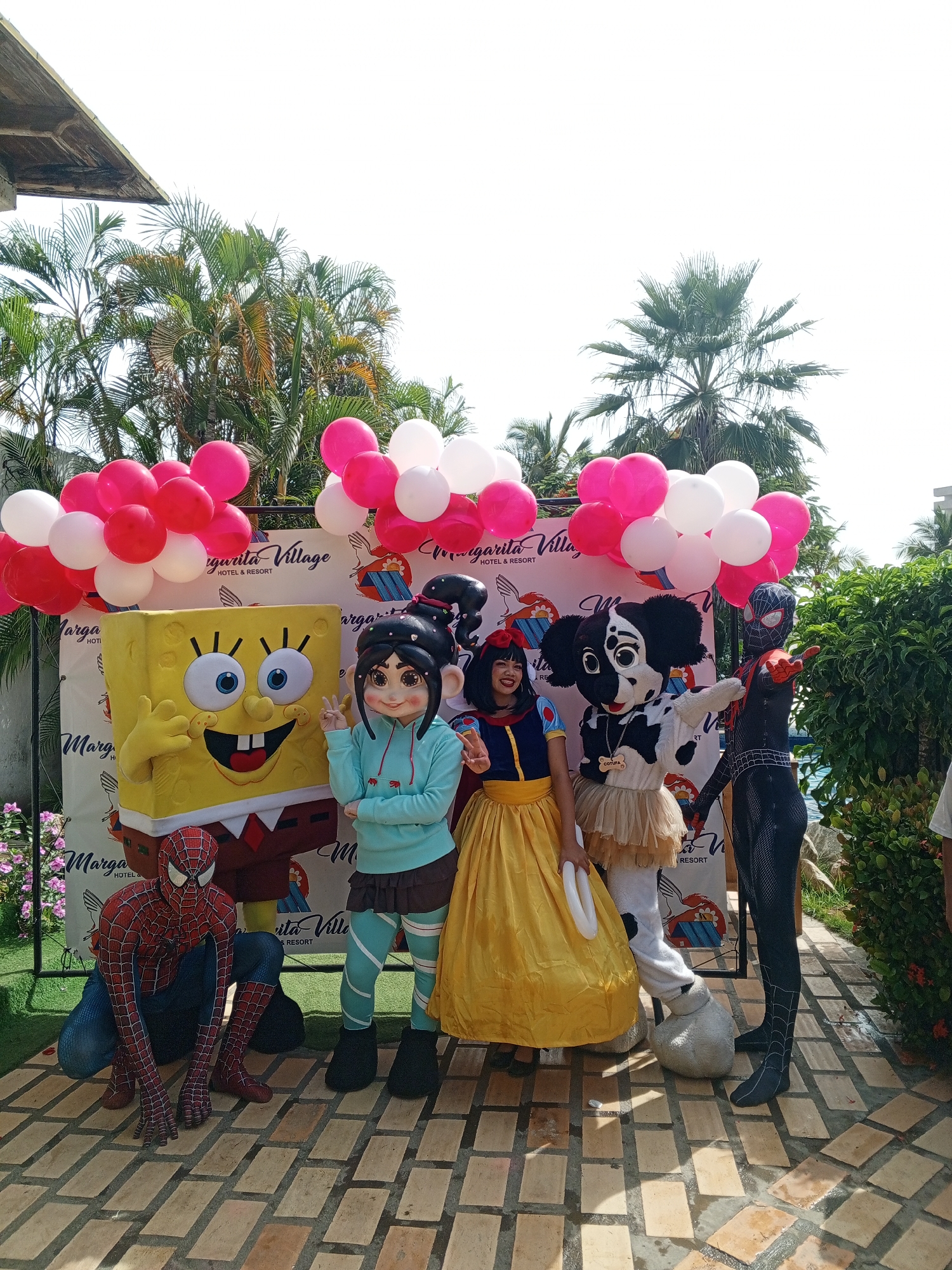 The image shows a group of people in costumes posing in front of a backdrop. The backdrop has the words "Margarita Village Hotel & Resort" printed on it and is decorated with pink and white balloons. The costumes include a Spongebob, Vanellope von Schweetz from Wreck-It Ralph, Snow White, a Dalmatian with a name tag reading "Cotufa", Spider-Man, and Miles Morales. The individuals are standing on a brick patio. In the background there is lush tropical foliage and a building. The location is likely in Porlamar, Venezuela, due to the name of the hotel on the backdrop.