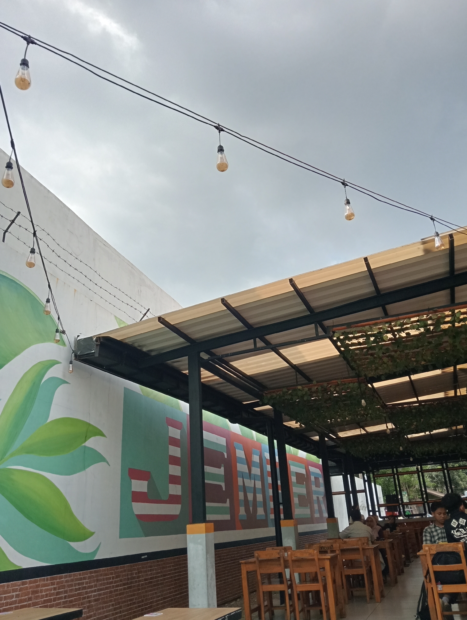 The image shows an outdoor seating area at what appears to be a cafe or restaurant in Jember, Indonesia. A large mural of green leaves and the word "JEMBER" in colorful block letters adorns the white wall on the left. Above, strings of Edison-style light bulbs are strung across wires against a cloudy sky. A corrugated metal roof, supported by dark metal beams and poles, provides shade over the seating area. The seating consists of wooden tables and chairs. In the background, several patrons are visible sitting at tables, some engaged in conversation. A black backpack is visible on the back of a chair in the foreground on the right. The foreground shows a close-up of a wooden table. The overall impression is a casual, open-air dining environment.