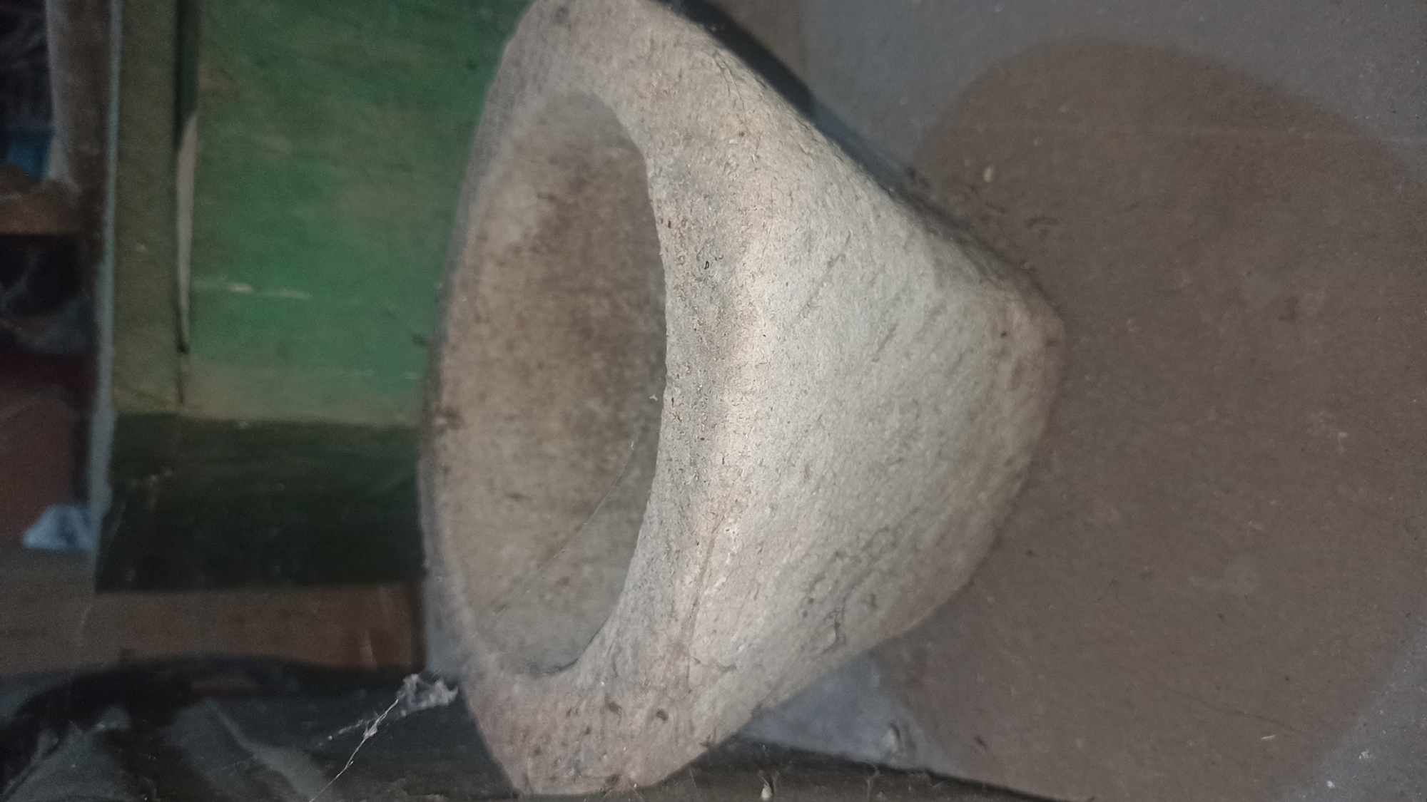 A close-up, eye-level, medium shot captures an old, rough-hewn stone mortar situated on a dark, dusty floor. The mortar is weathered and appears to be made from a coarse, light-colored stone, with an oval opening at the top. The exterior of the mortar tapers down to a broader base, and its surface is textured and uneven. A faint shadow is cast on the floor to the right of the mortar. In the background, a dark green, weathered wall and a dark, possibly fabric, object are visible, partially obscured by shadow. The overall impression is of an object in a storage or utility area, likely in an outdoor or semi-outdoor setting. The scene is devoid of any human presence or activity. The location is identified as Tuba, Indonesia.