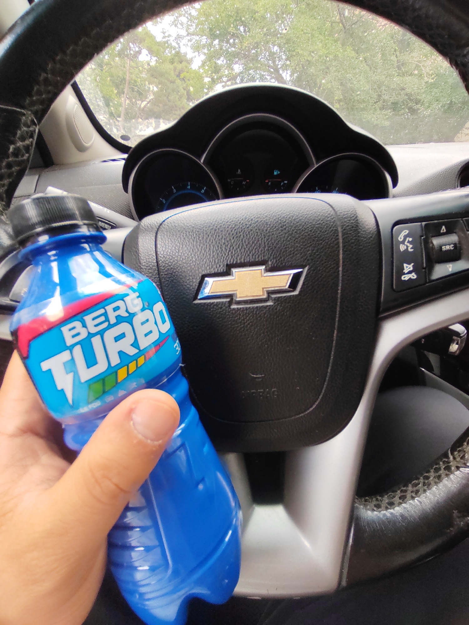 An eye-level, medium shot captures a hand holding a blue bottle of "BERG TURBO" energy drink in front of a car's steering wheel. The steering wheel, emblazoned with a Chevrolet logo, is black leather with textured grips on the sides. To the right of the steering wheel, control buttons are visible. The car's dashboard, featuring a speedometer and other gauges, is partially visible in the background. The scene is set inside a vehicle, with daylight and greenery visible through the windshield, suggesting an outdoor location, potentially in Baku, Azerbaijan, given the instruction. The hand holding the bottle appears to be a person's right hand, with the fingers wrapped around the bottle. The bottle itself is a bright blue plastic with a prominent "BERG TURBO" label in white and red lettering. The lighting suggests it is daytime.